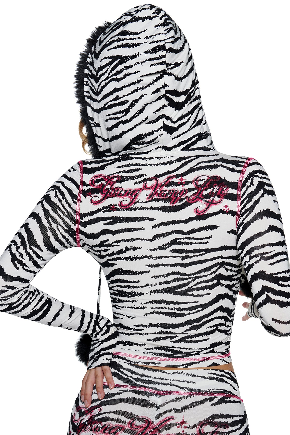 A person wearing an I.AM.GIA YVL BLARE FUR LINED HOODIE - ZEBRA, featuring a black fur-trimmed hood, pink contrast stitching, and the embroidered words “Young Vamp Life” on the back.
