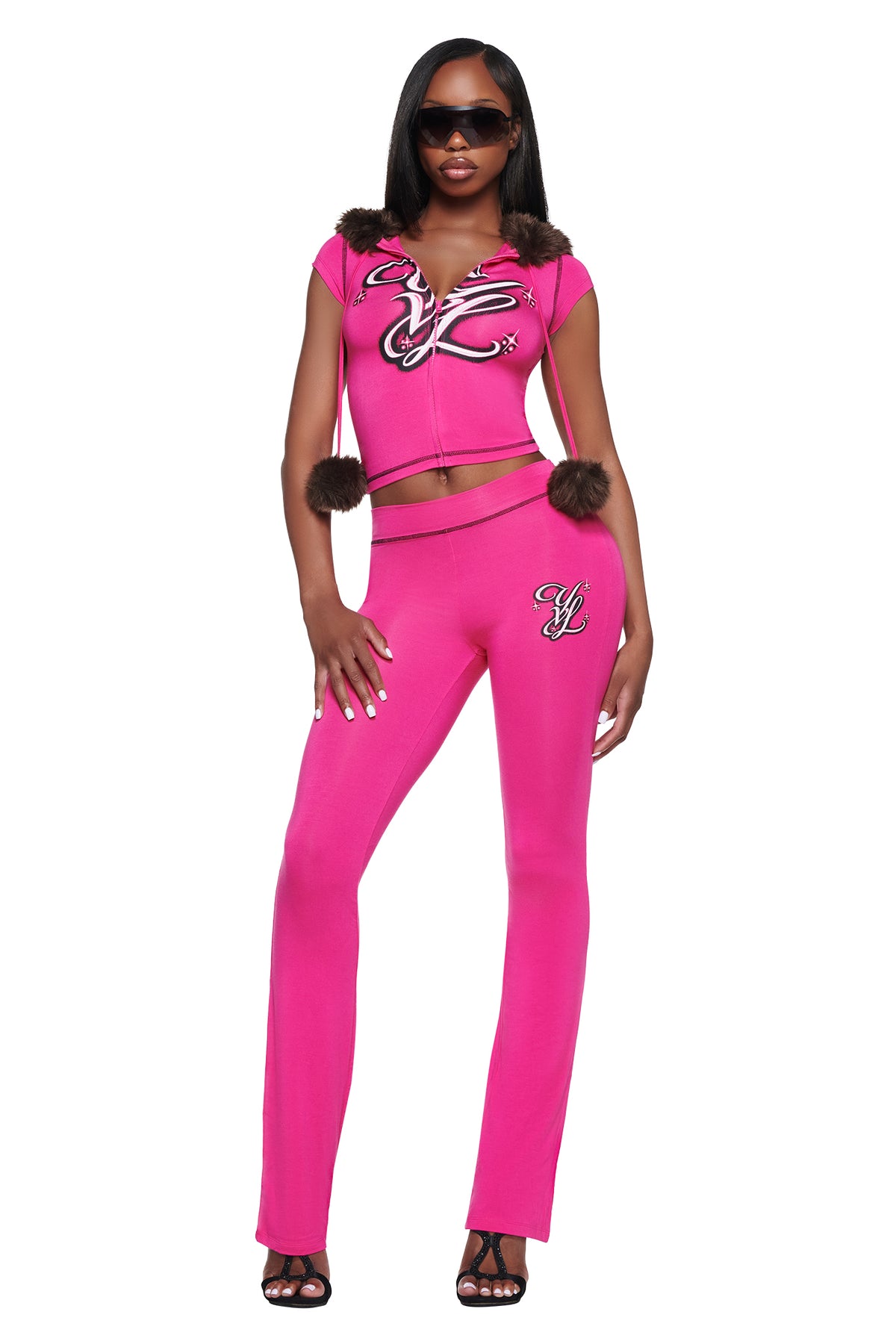 A woman wears a bright pink zip-up short-sleeve hoodie with brown faux fur trim and matching YVL BLARE TRACKPANT - PINK, both featuring black and white graphic logos.