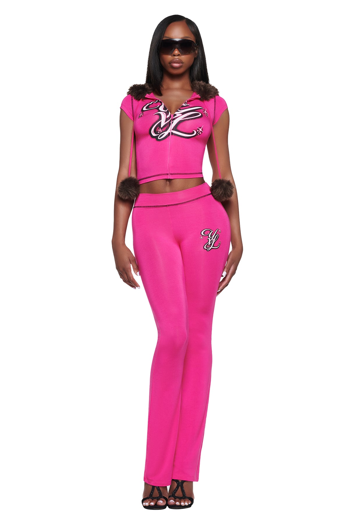 A person wears a matching hot pink I.AM.GIA YVL BLARE TRACKPANT - PINK set, consisting of a short-sleeved zip-up crop top with faux fur pom-poms on the hood strings and slim-fitting flare pants, both featuring embroidered logo detailing.