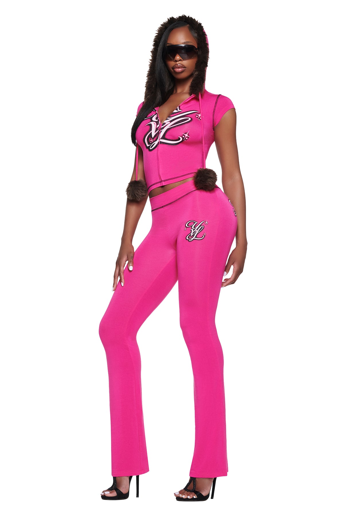 A person wearing a bright pink hooded short-sleeve zip-up top with faux fur trim and matching YVL BLARE TRACKPANT - PINK, both featuring embroidered logo detailing.