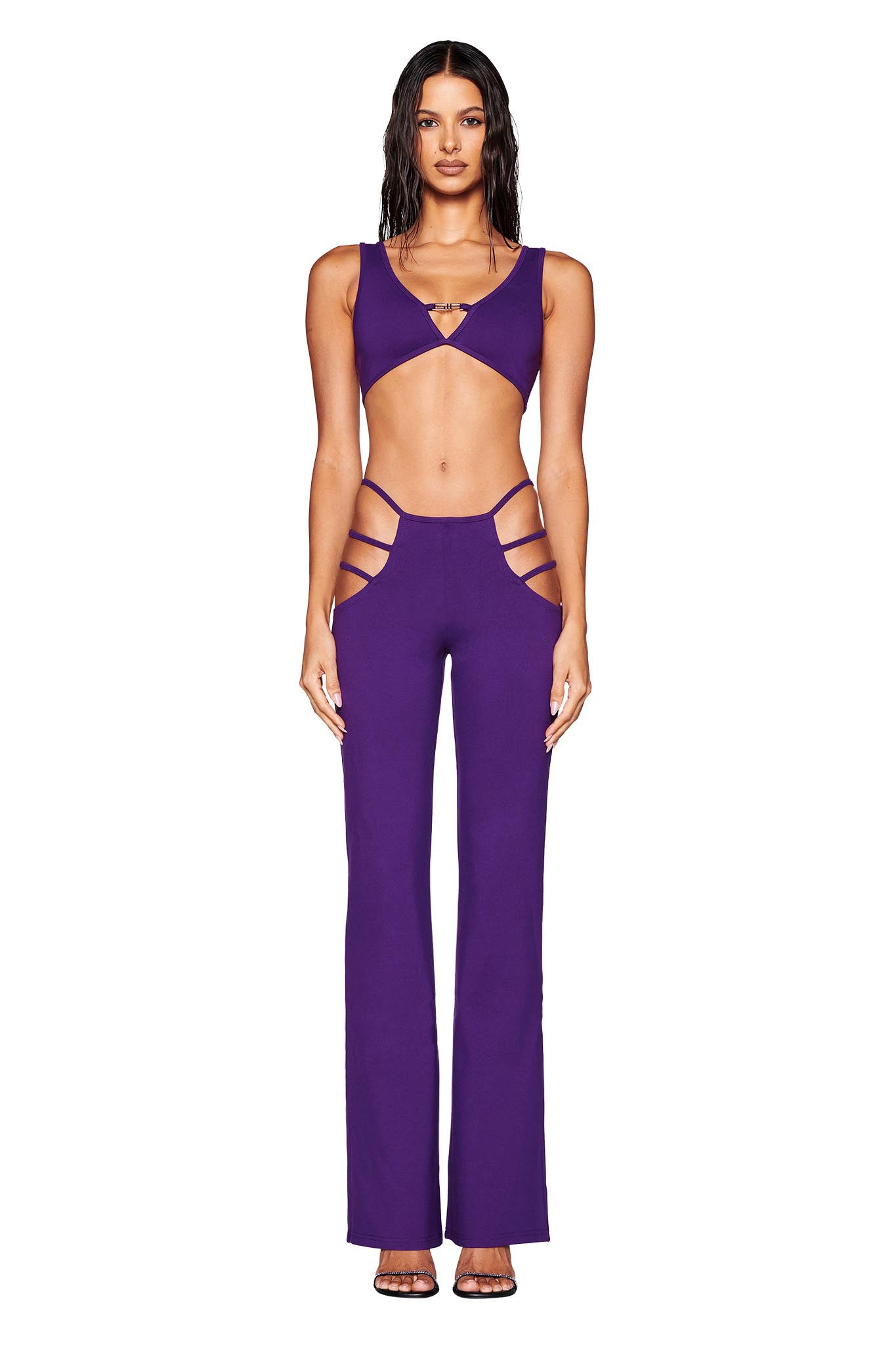 A person stands wearing the LUCID PANT - PURPLE from I.AM.GIA, reminiscent of a Euphoria Maddy outfit. The ensemble showcases a plunging neckline, strappy sides, and high-rise flared legs. The individual has long dark hair and is posing against a plain white background.