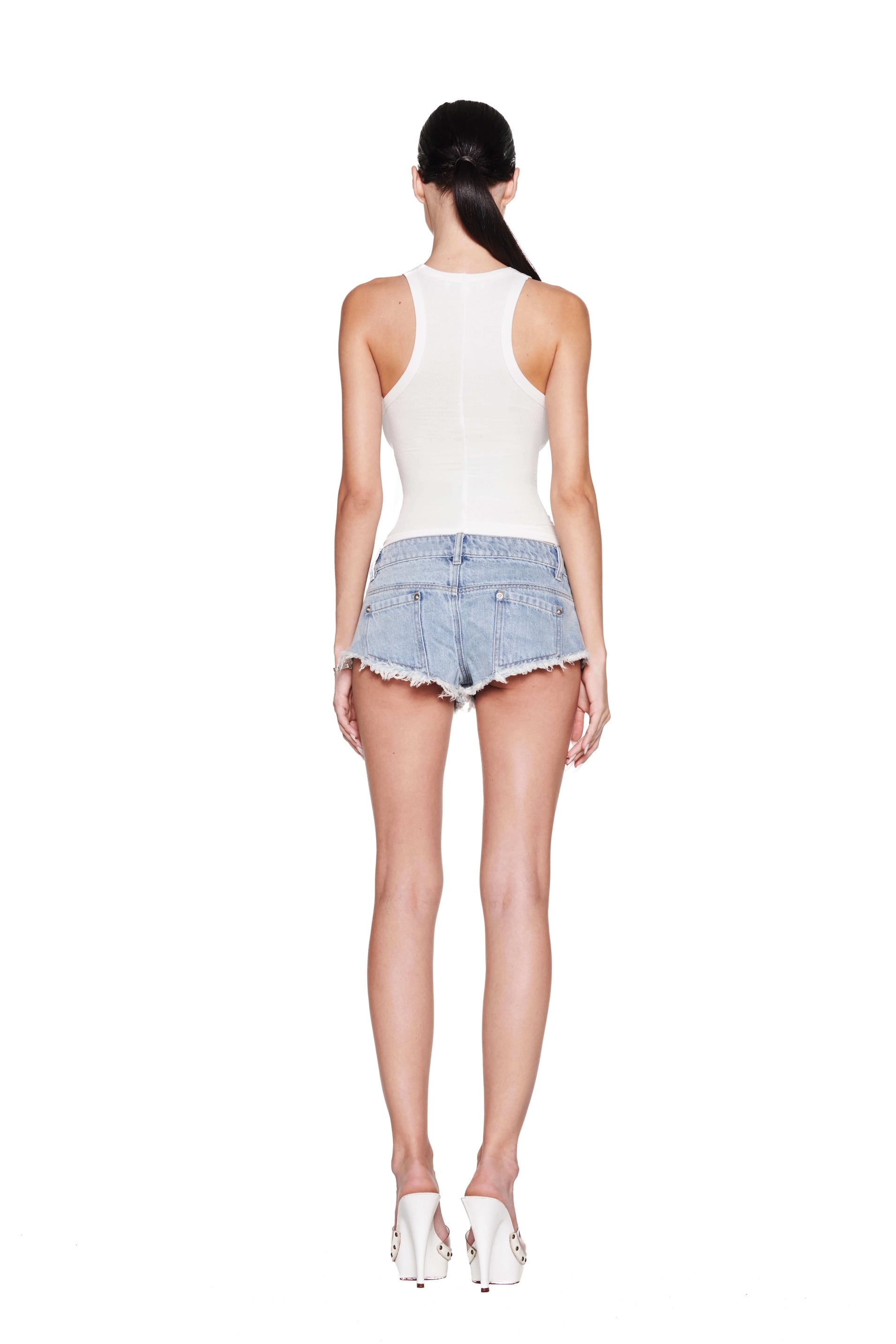 A person with long black hair is wearing an I.AM.GIA PENELOPE TANK - GIA GIRL in racer tank style, denim shorts, and white heels, standing with their back to the camera against a plain white background.