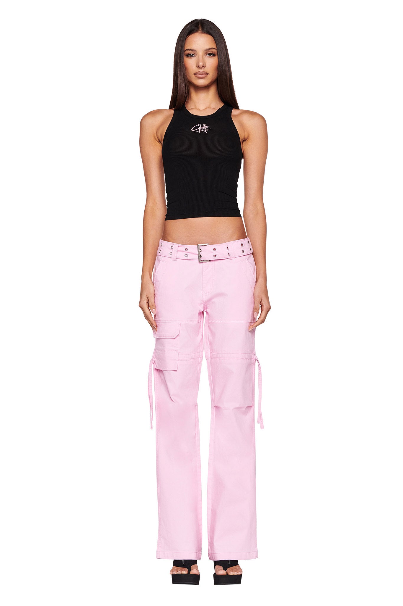 A woman stands facing the camera, wearing a black halter top with a graphic design from I.AM.GIA. She is also wearing the NAIMA CARGO PANT - PINK BABY from I.AM.GIA, featuring several pockets and ties, paired with black high-heeled sandals. She has long, straight hair. The background is plain white.