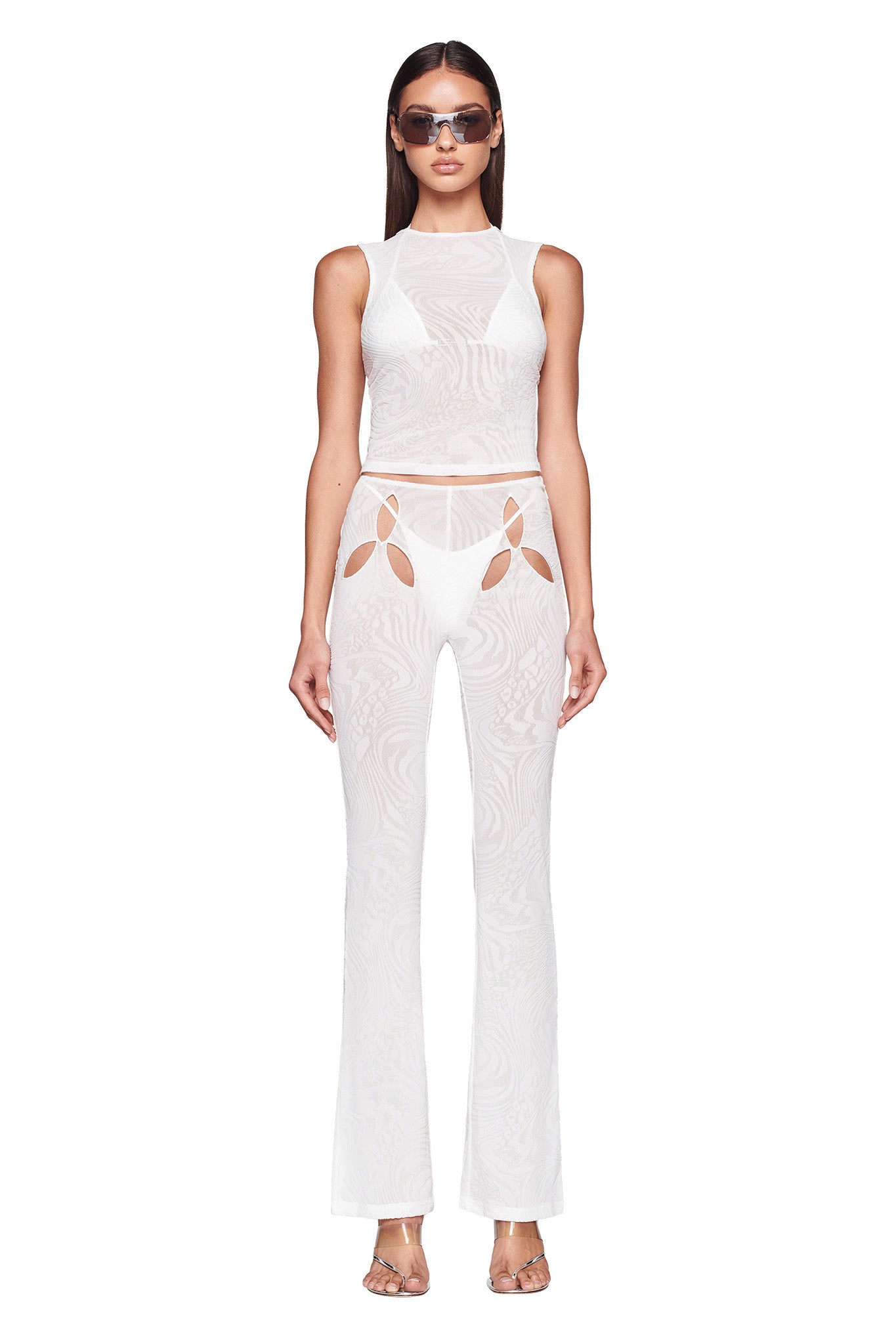 A person stands confidently, wearing a sheer white sleeveless top paired with the AMINA PANT - WHITE from I.AM.GIA, featuring unique cutout flower shapes. They also have on dark sunglasses and high-heeled sandals.