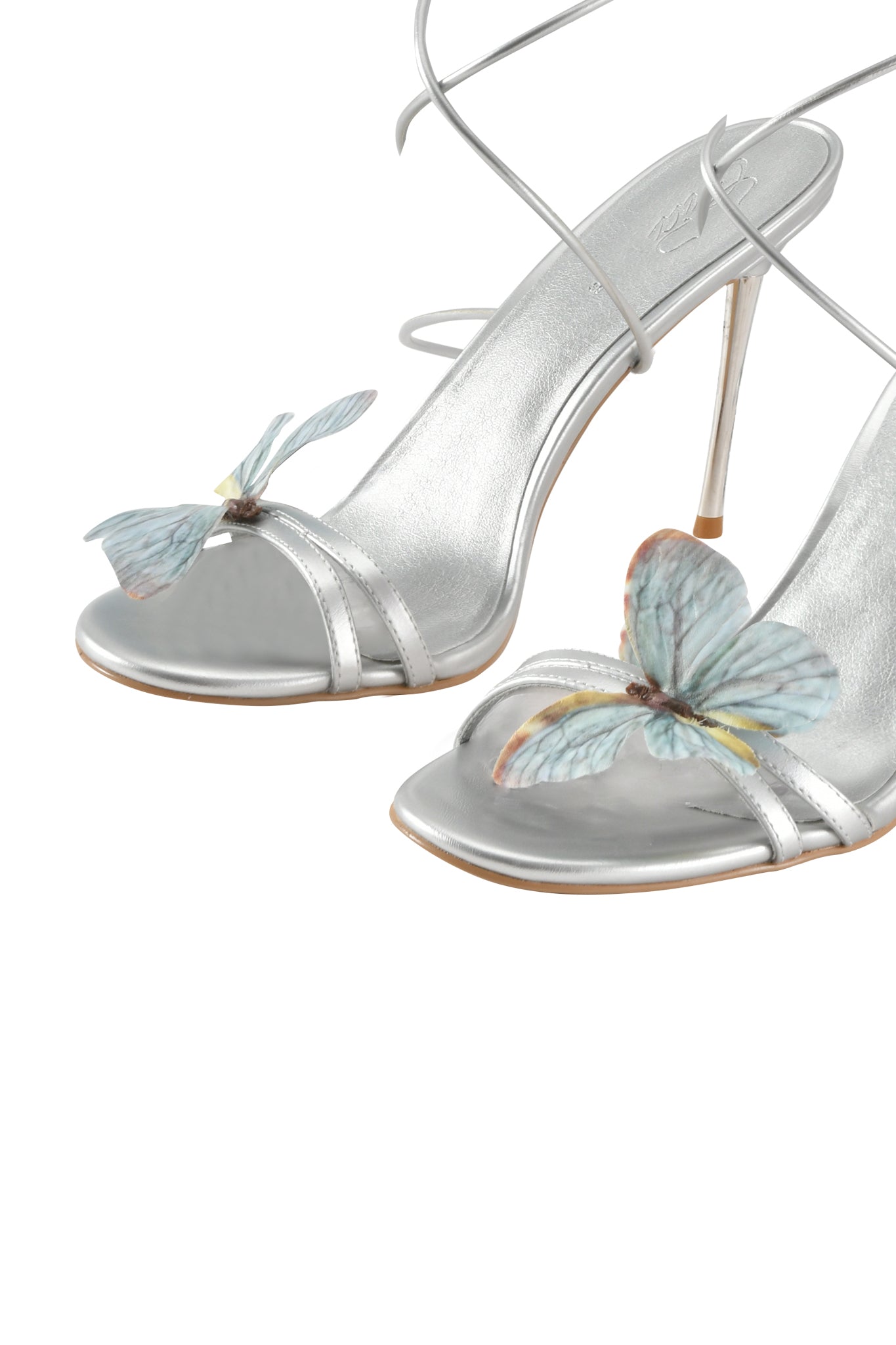 The SCARLETTE HEEL - SILVER by I.AM.GIA features elegant silver metallic strappy stiletto sandals with thin ankle straps and blue butterfly appliques on the open toe, set against a white background.