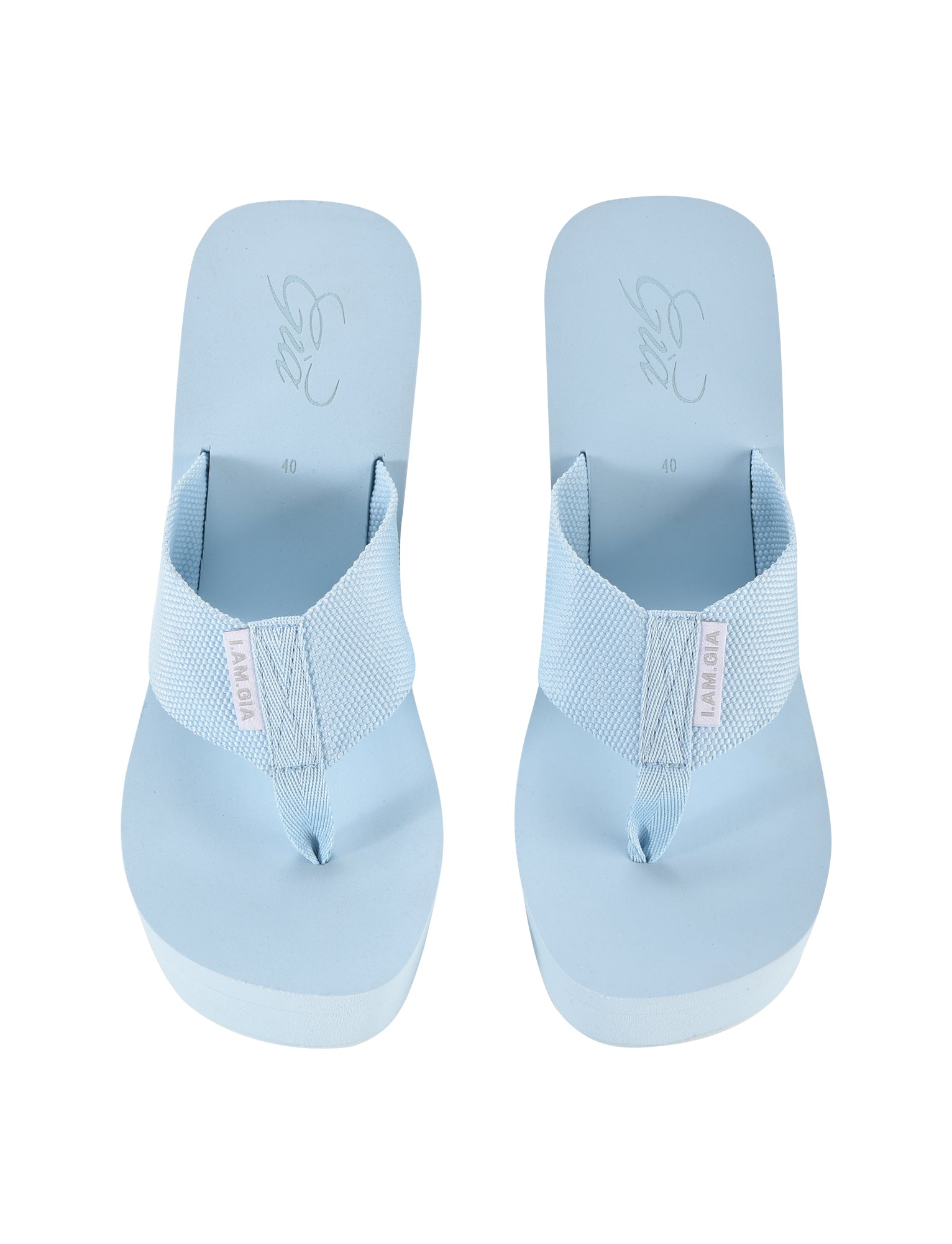 A pair of baby blue flip-flops featuring wide fabric straps and thick soles. The brand name "I.AM.GIA" is visible on a small label attached to each strap, adding a touch of luxury. These SHELBY FLIP FLOPS have a minimalist design and are positioned side by side, viewed from above.