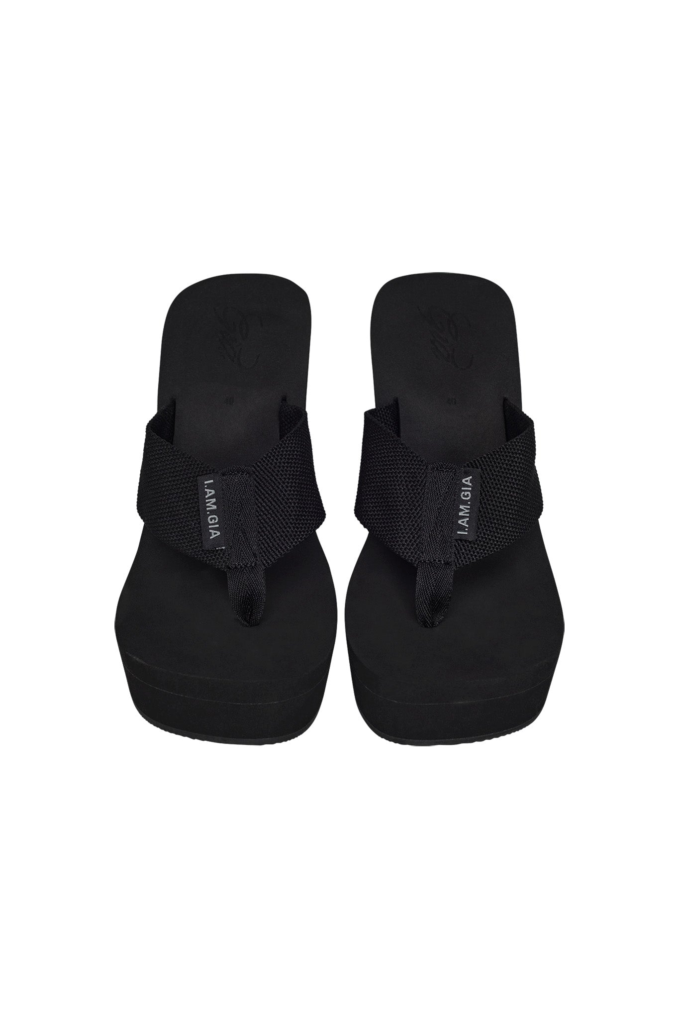 The SHELBY FLIP FLOP - BLACK from I.AM.GIA is a pair of black platform flip-flops featuring fabric straps and a fabric thong design. The footbeds have a slight texture, and the left strap has the "I.AM.GIA Malibu" label sewn on the outer side. Both flip-flops have thick soles for added height.