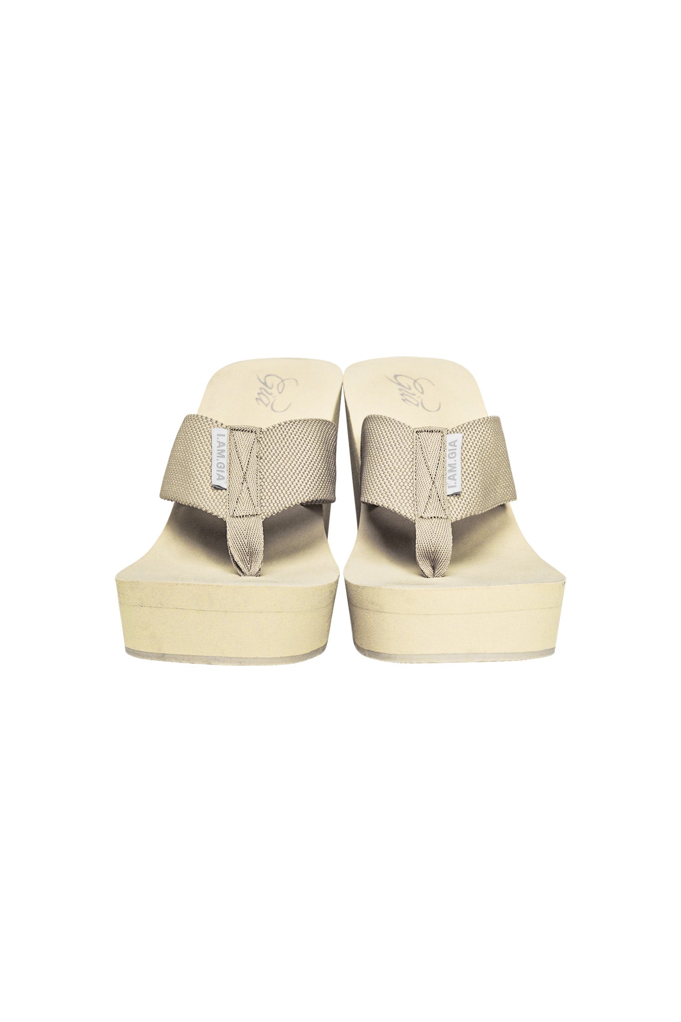 A pair of SHELBY FLIP FLOP sandals in Neutral Tan by I.AM.GIA, featuring wide fabric straps with a small I.AM.GIA brand tag on each strap. The platform flip-flops have a slightly textured sole and are set against a plain white background.