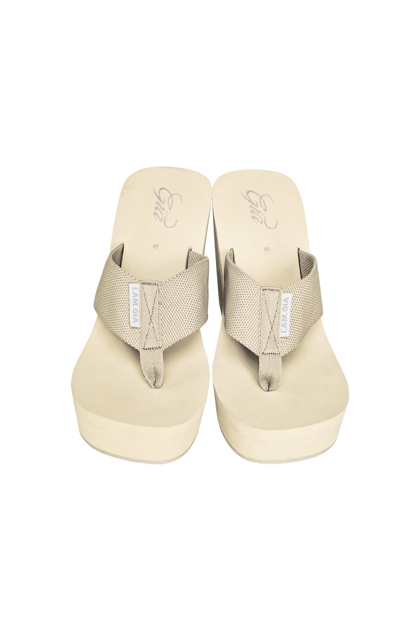 A pair of SHELBY FLIP FLOP - NEUTRAL : TAN by I.AM.GIA, featuring wide fabric straps and white I.AM.GIA labels. The textured straps cross over the top, forming a comfortable footbed, with the brand logo visible on each sandal's insole.