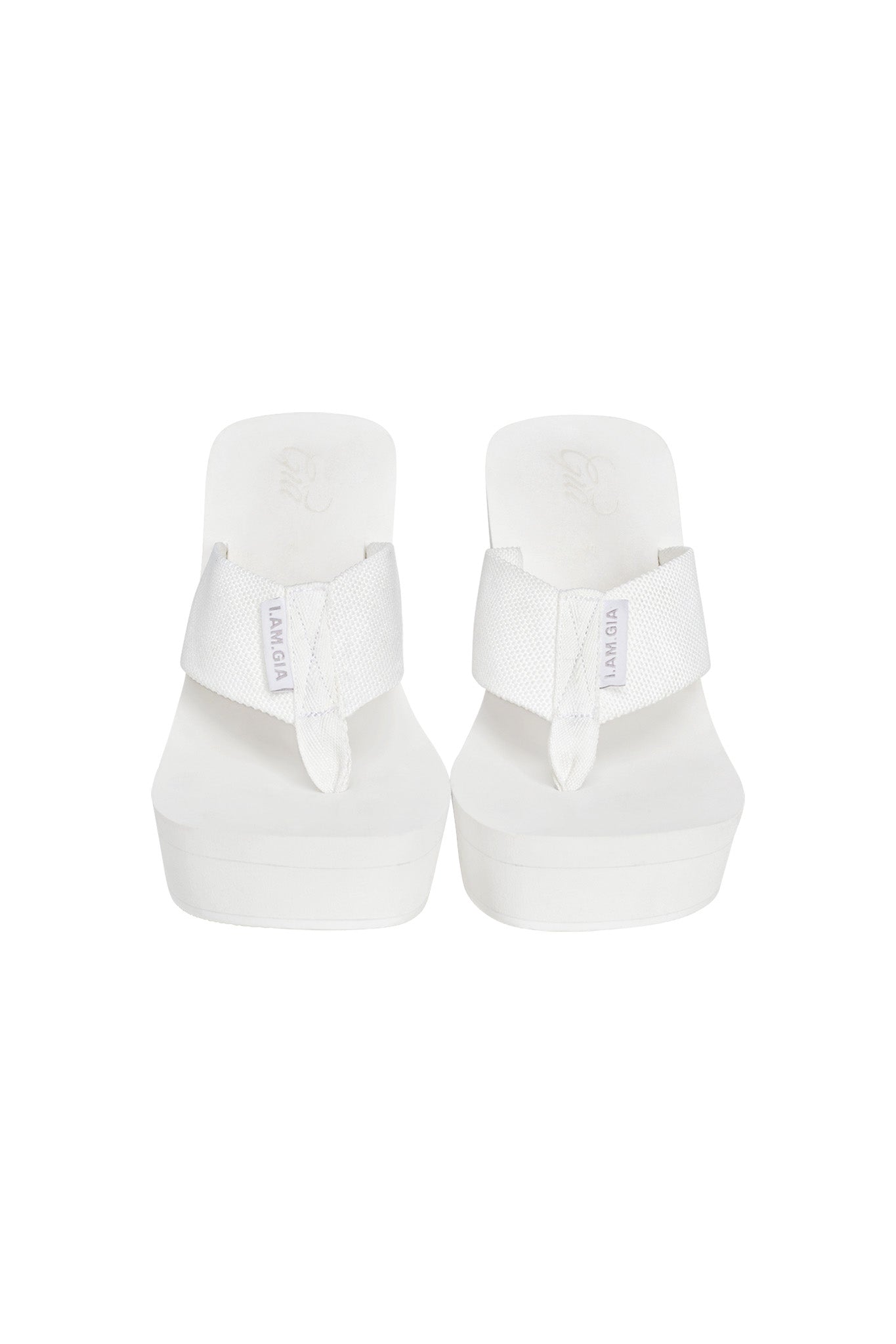 The SHELBY FLIP FLOP - WHITE by I.AM.GIA is a pair of white, open-toe platform wedge sandals featuring wide, padded straps across the top. The brand name is visible on a small tag attached to the left strap. Designed for comfortable wear, these sandals offer a simple and minimalist look.