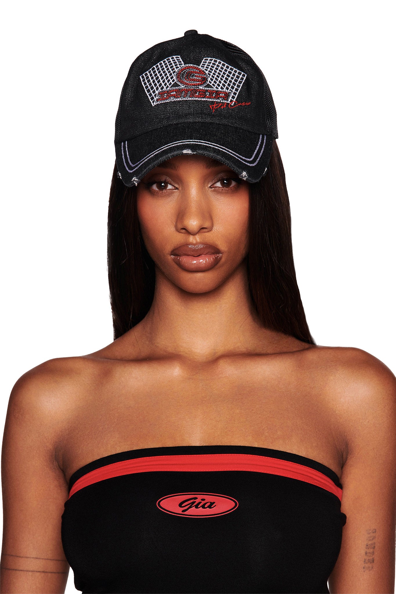 A person with long straight hair is wearing the KIMANI CAP in black from I.AM.GIA, featuring red and white designs and text on the front. They are dressed in a black strapless top with red stripes and an "I.AM.GIA" logo. The background is plain white.