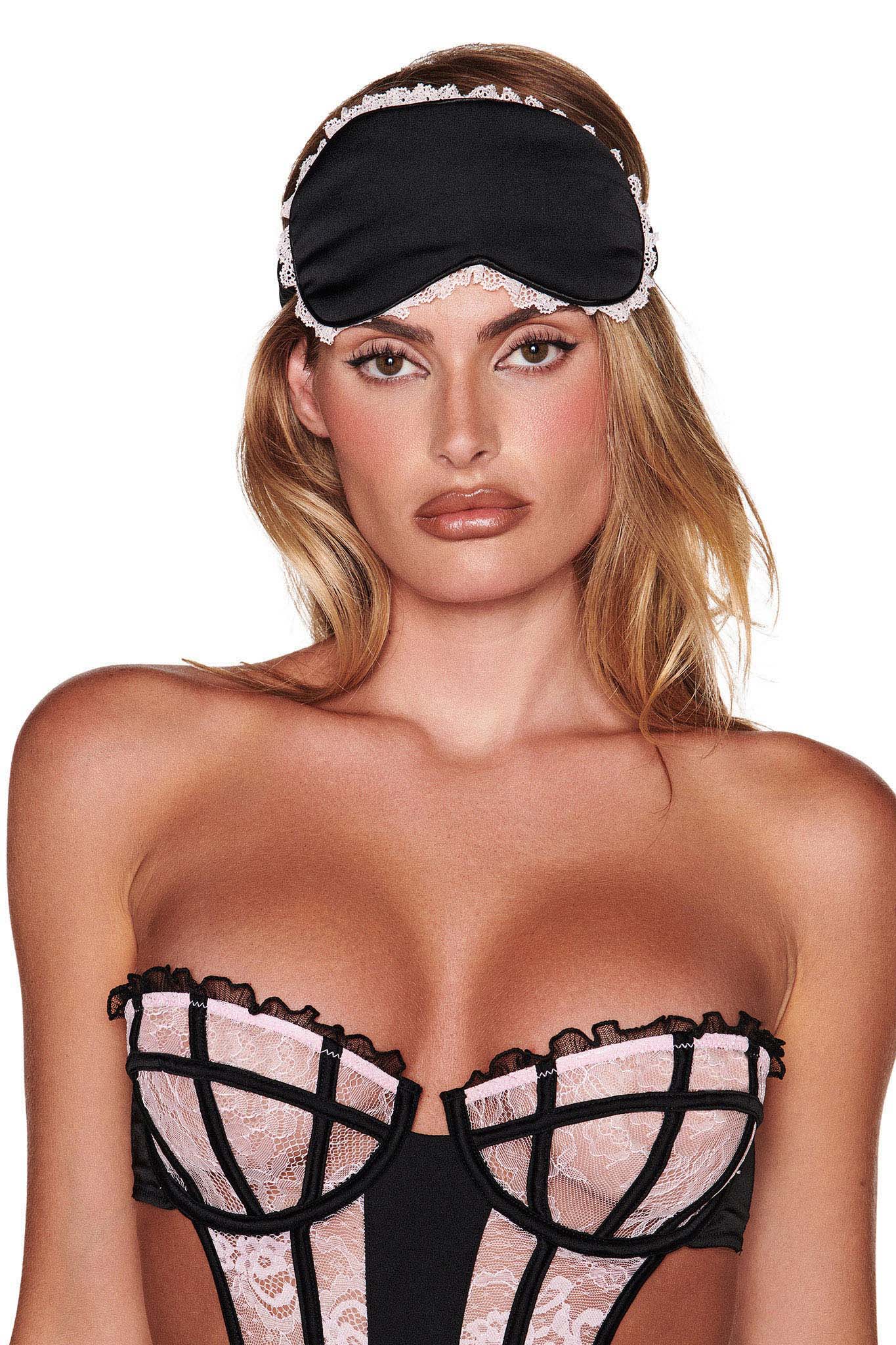 A person with long hair wears the MARILYN EYE MASK in black by I.AM.GIA, paired with a strapless corset-style top featuring light pink and black lace adorned with delicate pink ruffles. The background is plain white.