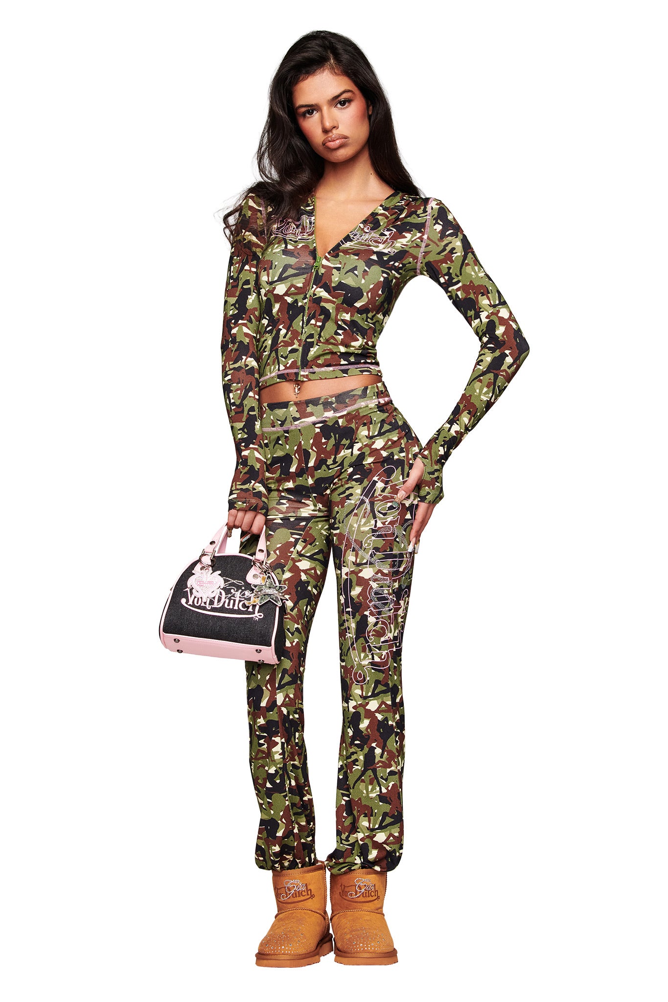 A woman stands against a white background in the I.AM.GIA X VON DUTCH BLARE HOODIE - GIA GIRL CAMO, paired with matching pants, a black and pink Von Dutch bag, and tan Timberland-style boots.