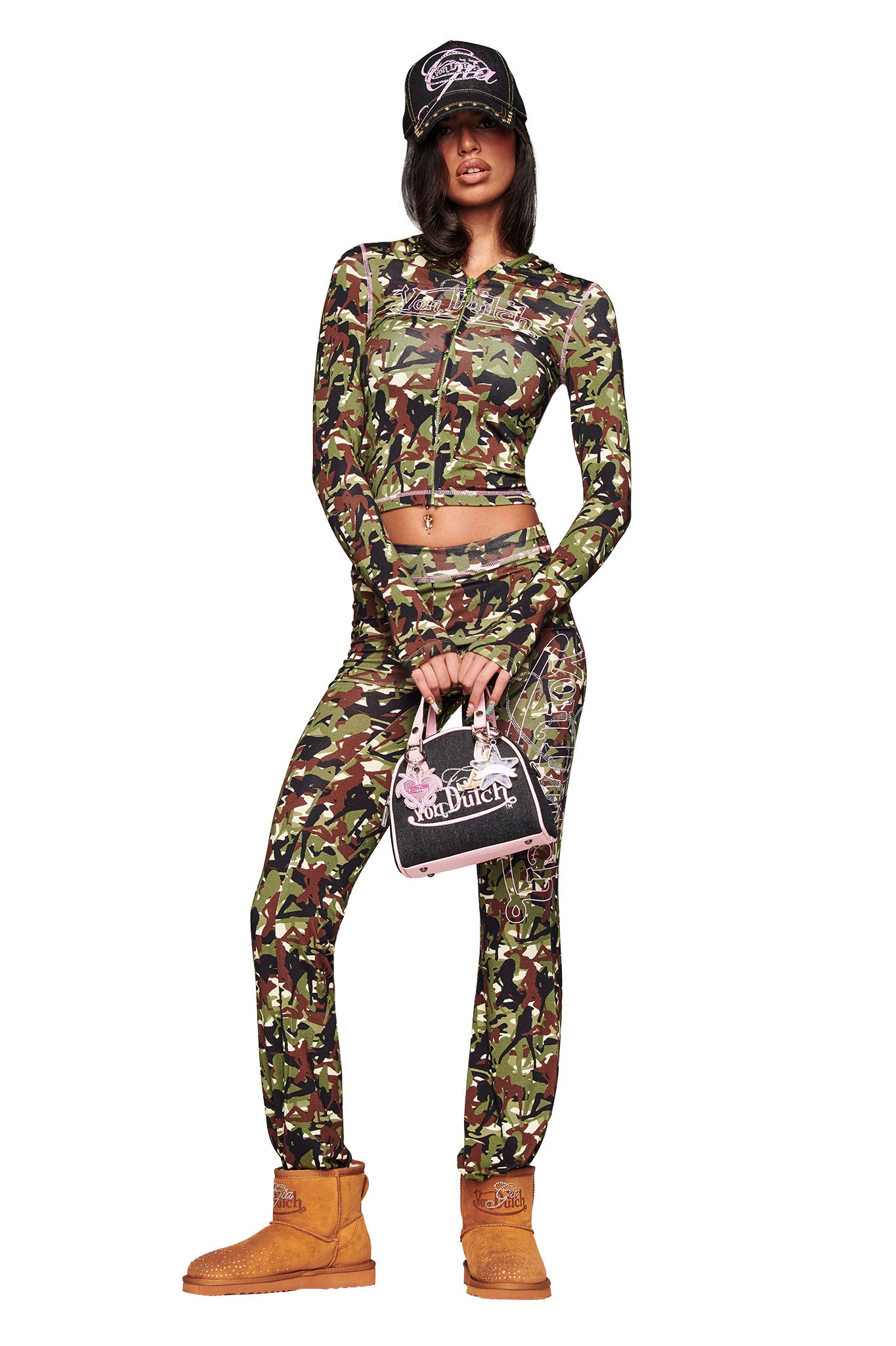 A person poses confidently in a green camo crop top and pants, wearing the I.AM.GIA X VON DUTCH FAUX SHEARLING BOOT in brown and a black cap, and holding a small black and pink Von Dutch handbag against a white background.