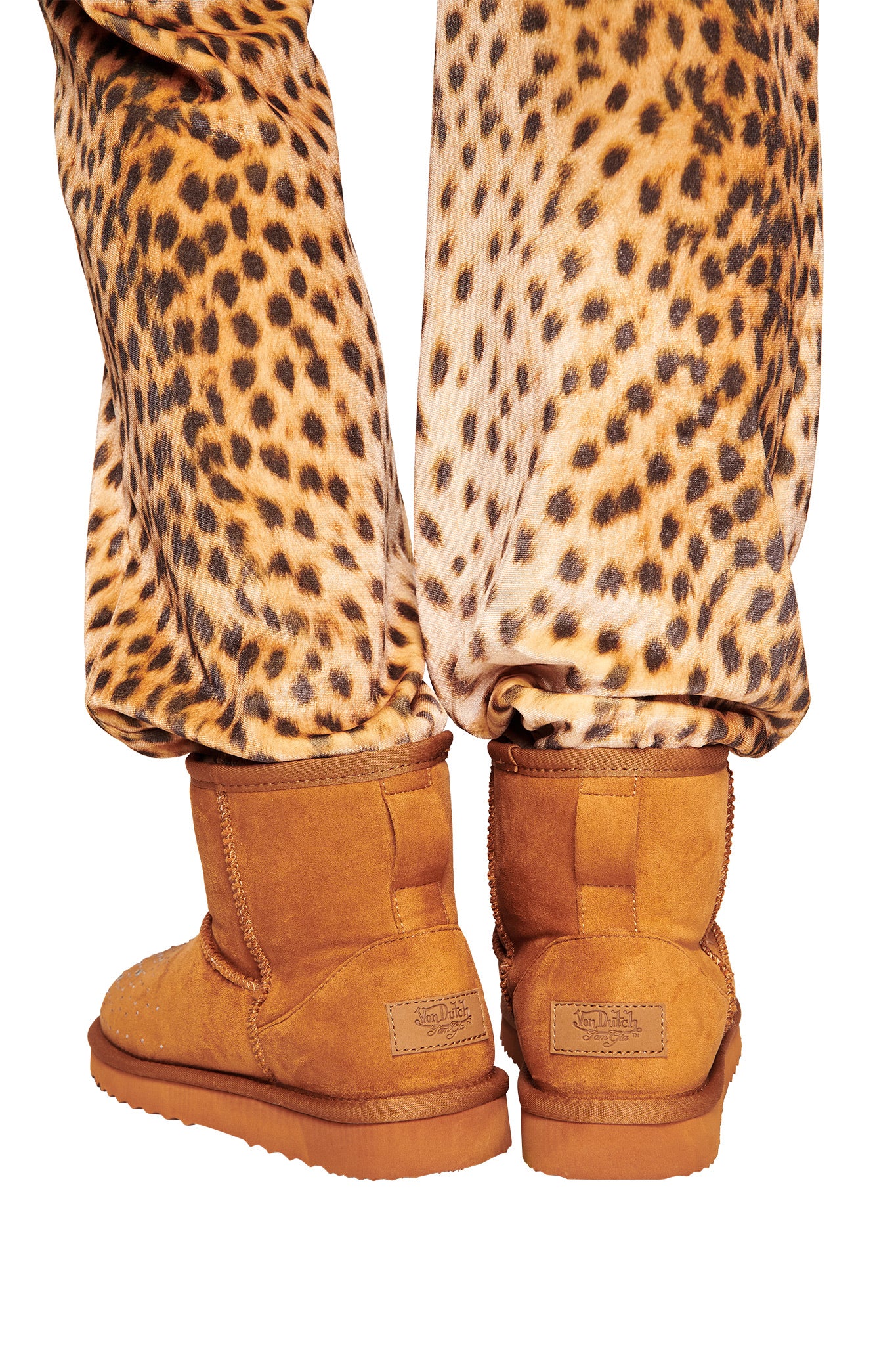 A person stands with legs together in loose, bold leopard print pants and I.AM.GIA X VON DUTCH FAUX SHEARLING BOOT - BROWN, viewed from behind to highlight only the lower legs and feet.