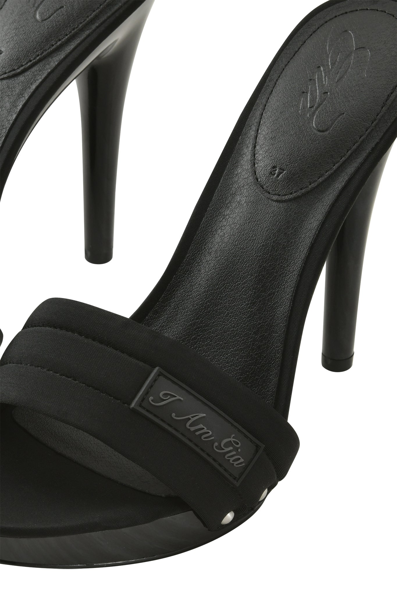 A close-up of the NICKY HEEL - BLACK NEOPRENE by I.AM.GIA, featuring black slip-on mule sandals with a padded vamp strap adorned with a metal “I Am Gia” plate and sleek, thin black stiletto heels for an elegant finish.