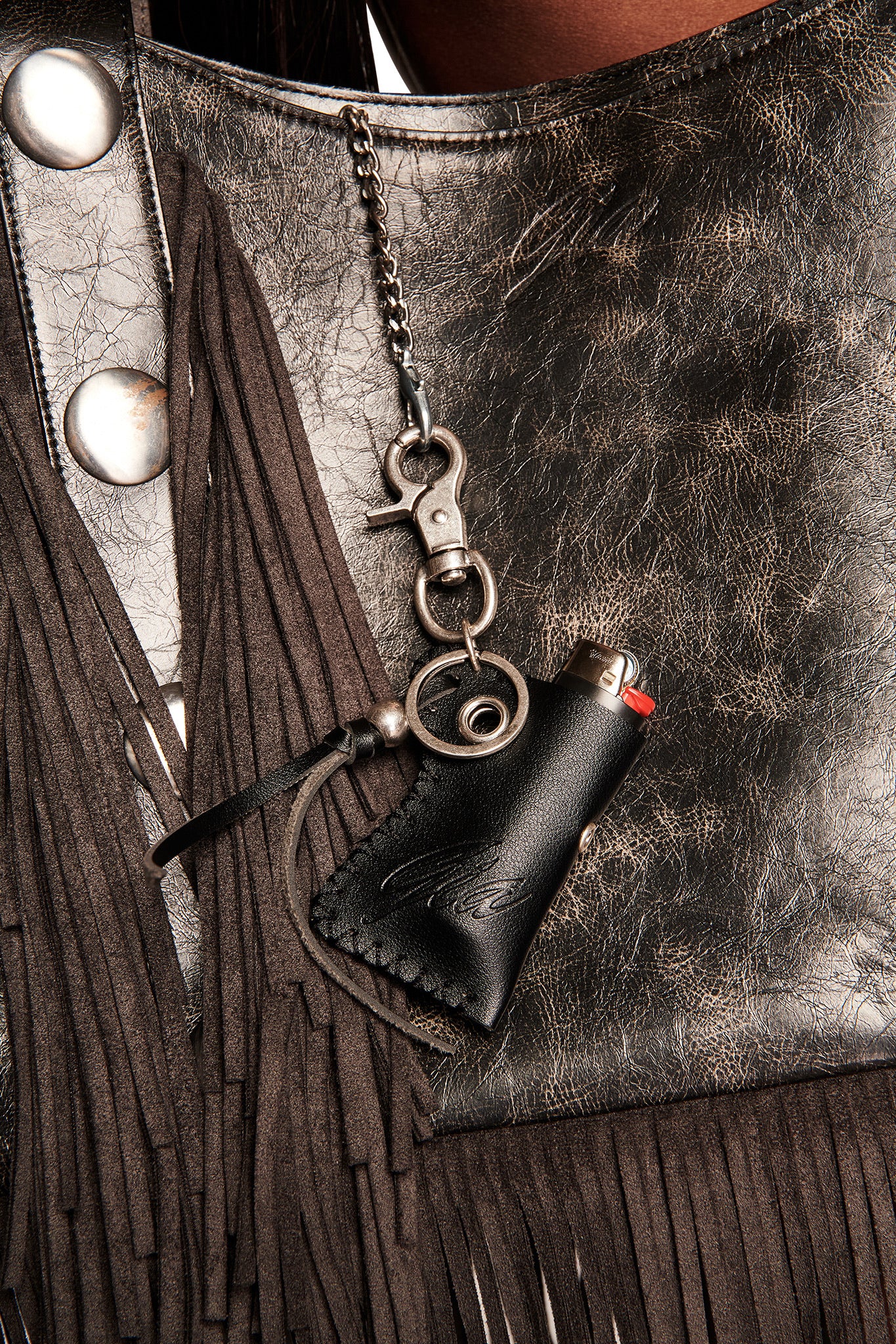 A close-up of a metallic, textured leather bag with fringe details features the I.AM.GIA VENUS LIGHTER KEYCHAIN - BLACK, a black faux leather lighter holder with metal clasp and red lighter, hanging by a chain like a keychain.