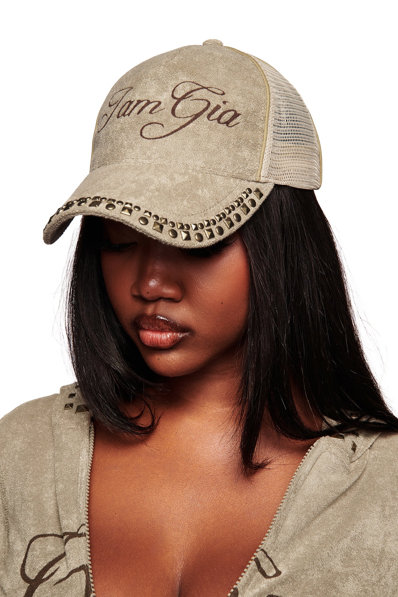 A woman wears the I.AM.GIA ISSHA TERRY HAT in Stone, featuring "I am Gia" embroidery and studded brim accents, paired with a matching beige zip-up top. She looks downward, her face partially shaded by the hat.