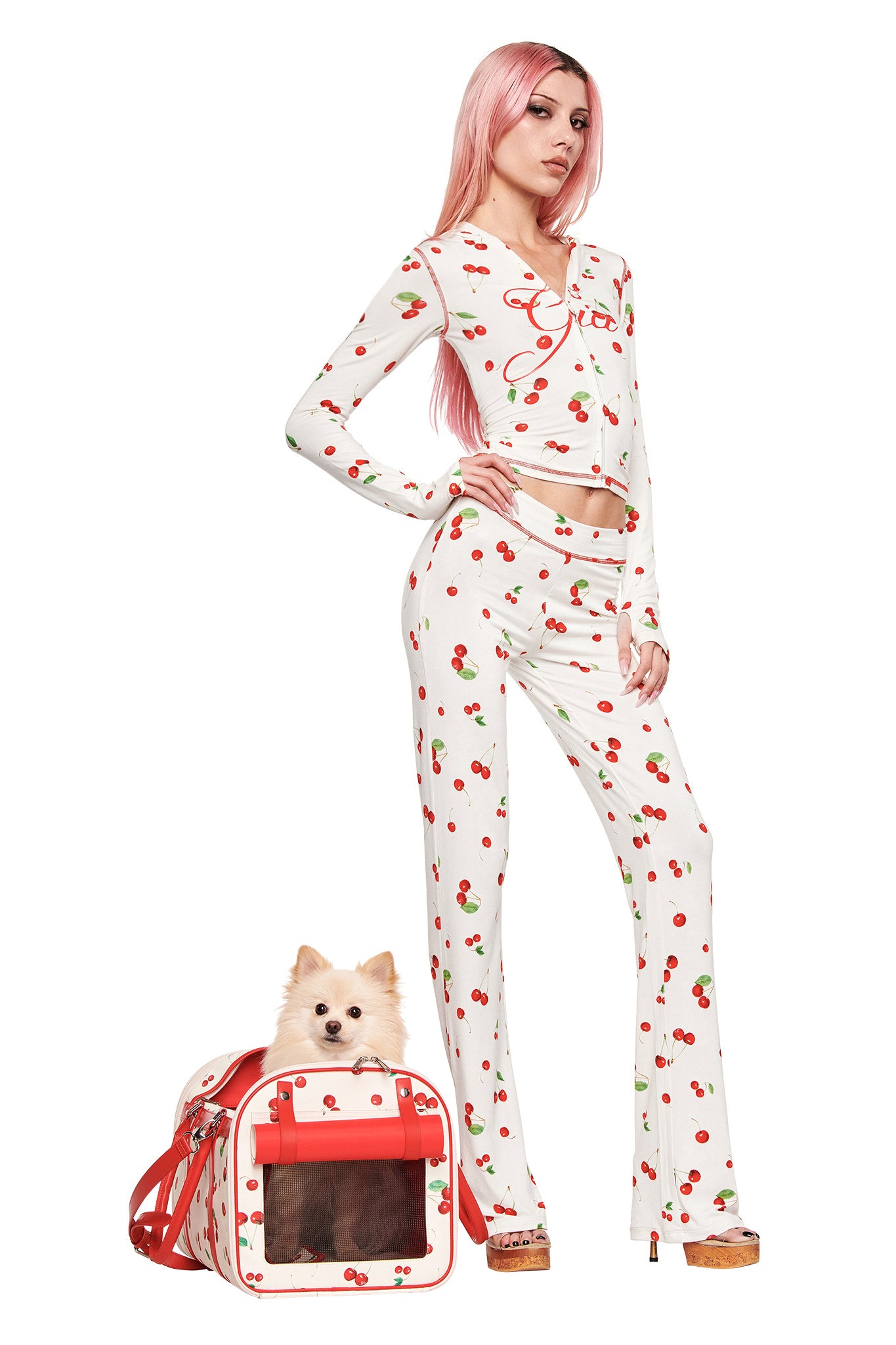 A woman with long pink hair wears a white pajama set with the Cherry on Top print, standing beside a fluffy dog sitting in the matching I.AM.GIA TRIXX PET CARRIER - CHERRY ON TOP. The background is plain white.