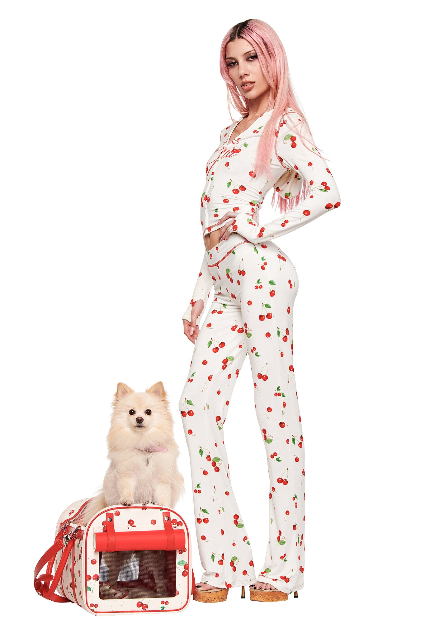 A woman with long pink hair wears a white Cherry on Top outfit by I.AM.GIA, posing confidently beside her small white fluffy dog and the matching I.AM.GIA TRIXX PET CARRIER - CHERRY ON TOP, set against a plain white background.