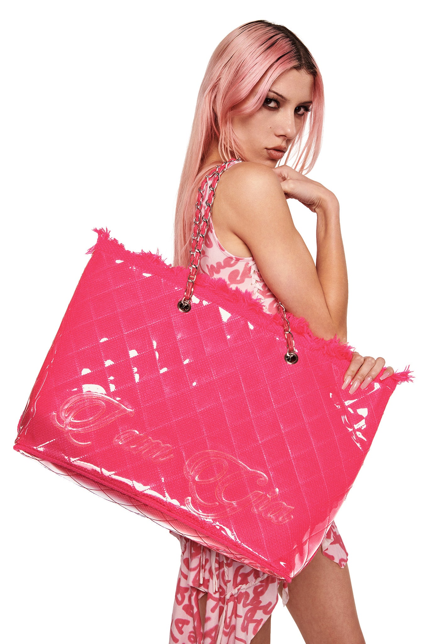 A woman with long pink hair poses holding the I.AM.GIA DANNI TOTE BAG in hot pink, featuring metal chain handles. She wears a pink and white patterned outfit against a white background.