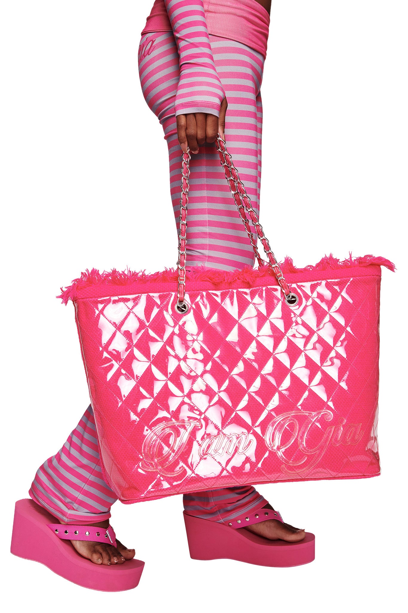A person in pink and gray stripes with pink platform sandals carries the I.AM.GIA DANNI TOTE BAG in hot pink, featuring chain handles, frayed edges, and a stylish PVC shell.