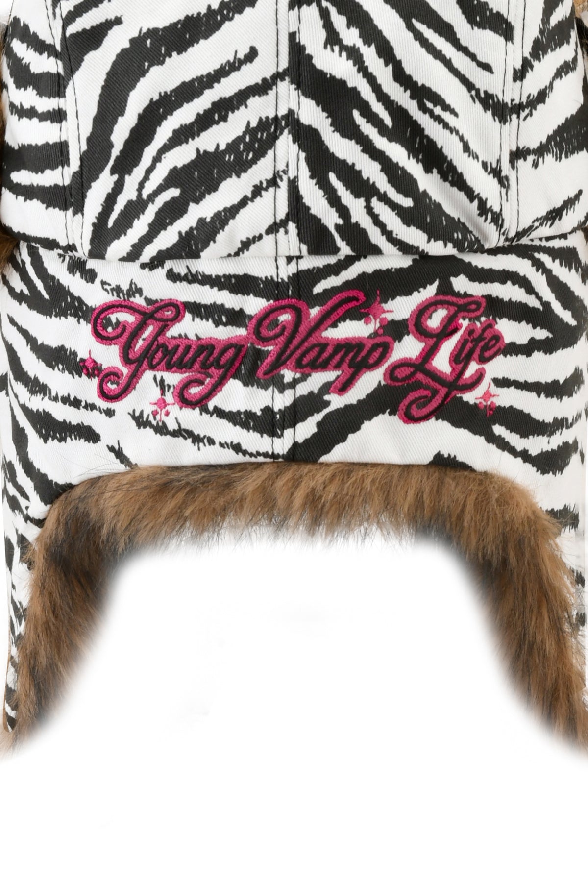 YVL FUR TRAPPER HAT - ZEBRA in a black-and-white print with brown faux fur trim and “Young Vamp Life” embroidered in pink script.