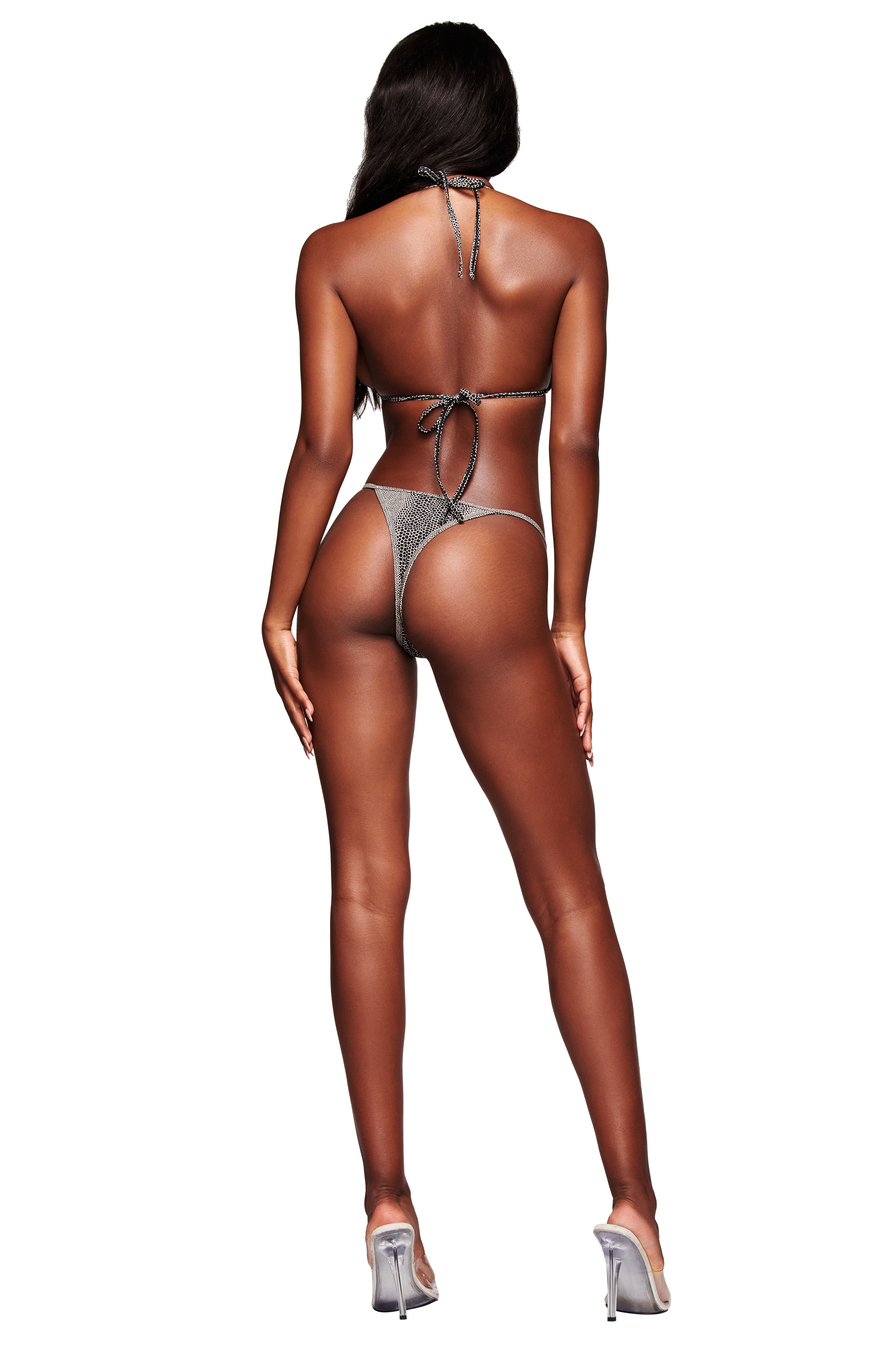 A woman with long dark hair faces away, wearing the I.AM.GIA AVA BIKINI TOP - SERPICO, a sparkling silver triangle bikini top, paired with clear high-heeled shoes against a white background.