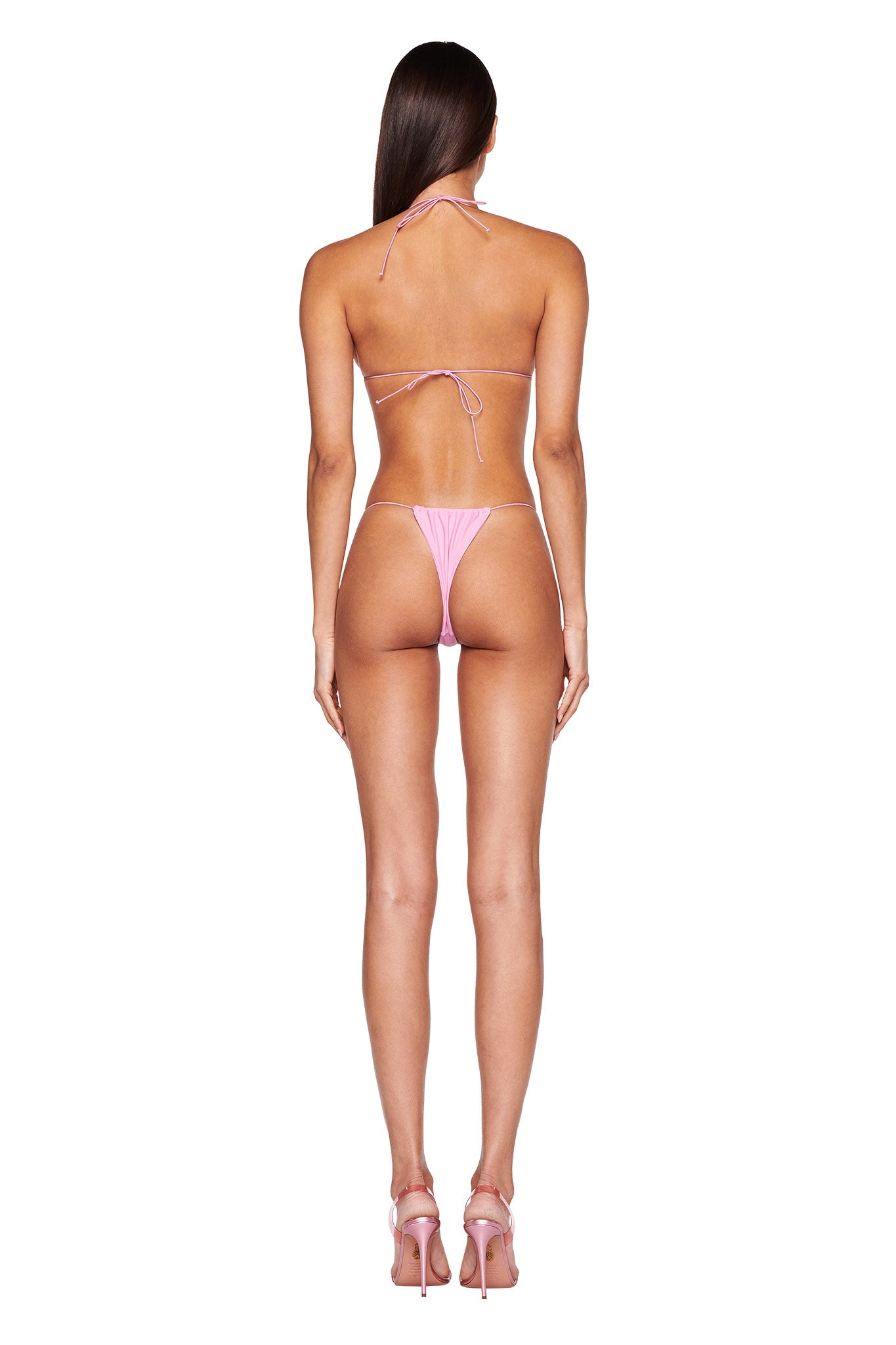 A person is standing facing away from the camera, wearing the BAMBI BIKINI TOP in pink by I.AM.GIA and a pair of high-heeled shoes. The background is plain white, highlighting their long, straight dark hair. This chic ensemble perfectly captures the edgy essence of I.AM.GIA.