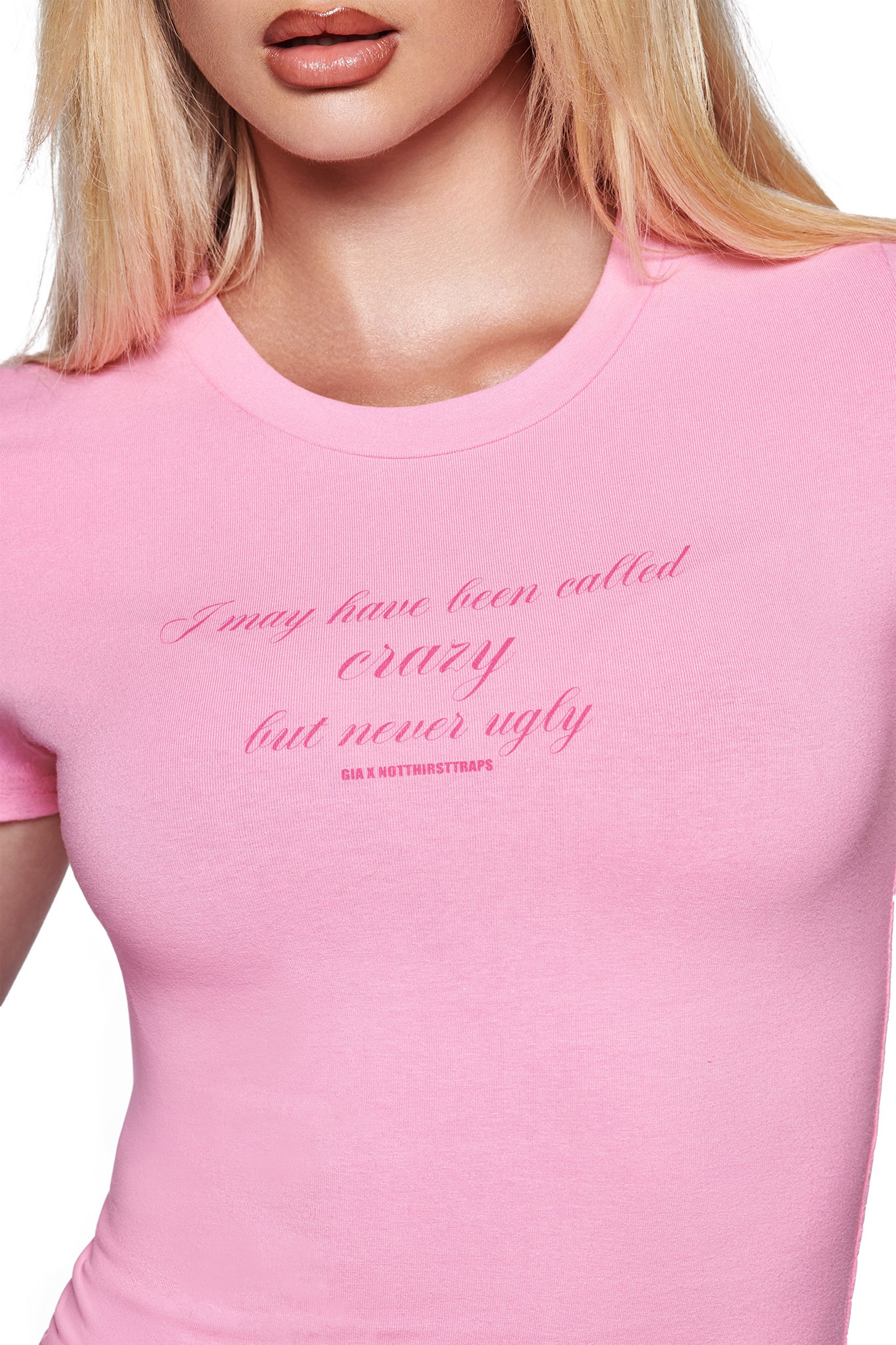 A woman with light skin and long blonde hair wears the I.AM.GIA POLINA CRAZY TEE in pink, a fitted soft cotton jersey top with “I may have been called curvy but never ugly” in pink cursive. Only her torso and lower face are visible.