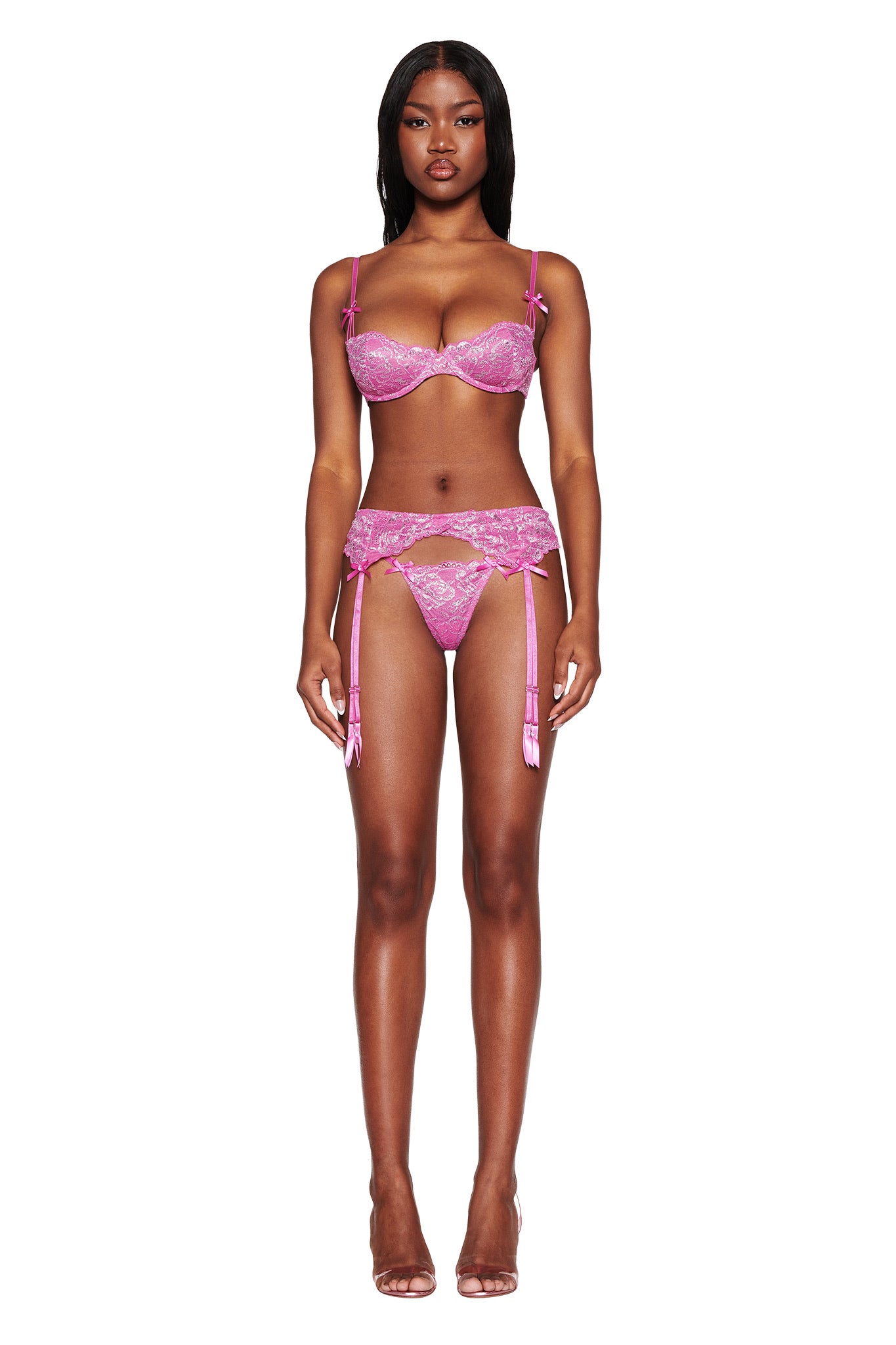 Against a plain white background, the woman elegantly wears the RAINE 3 PIECE METALLIC SET in pink by I.AM.GIA, a Y2K-inspired lace lingerie set with garter straps. Her metallic magenta high heels perfectly complement her stylish ensemble.