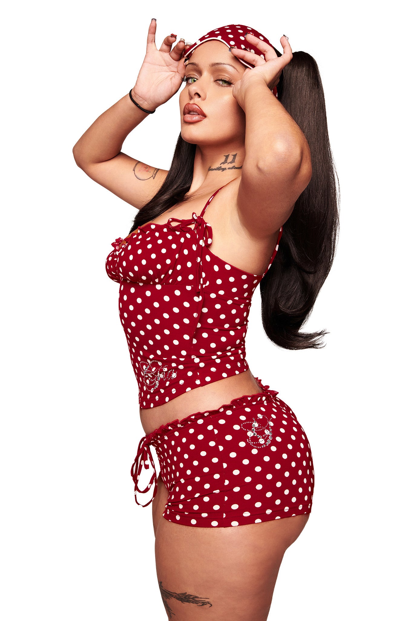 A woman models the I.AM.GIA MELANIE CAMI in red polka dot with adjustable straps and matching shorts, her long dark hair and visible arm and thigh tattoos adding to her confident look.