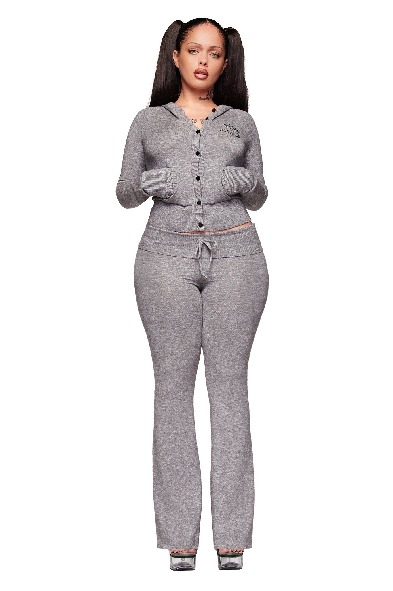 A woman with long, dark pigtails stands against a white background, hands in her pockets, wearing I.AM.GIA's JESSA PANT - GREY with a fitted light gray cardigan and heeled sandals.