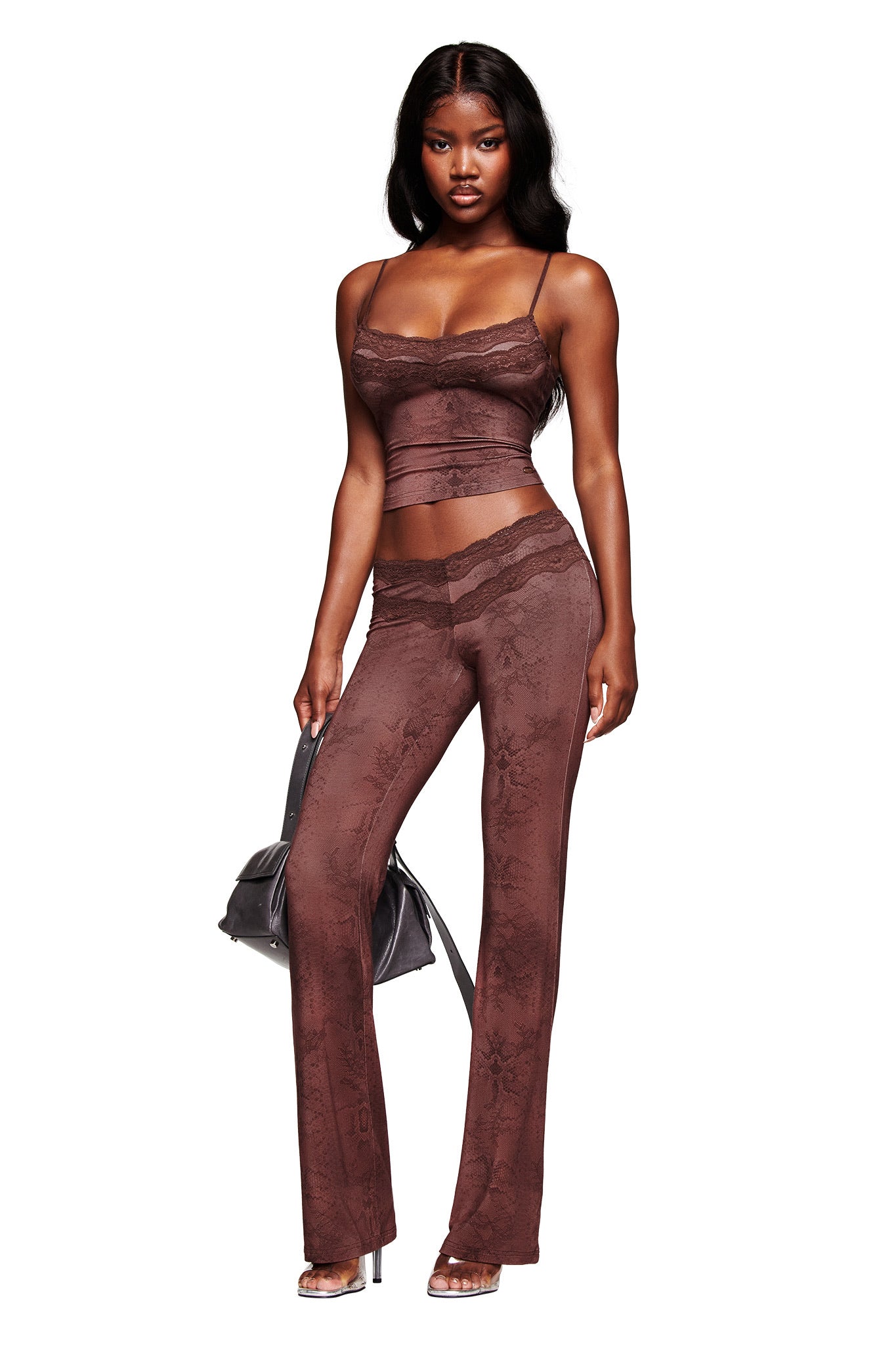 A woman stands confidently against a white background wearing the I.AM.GIA BEAU TOP in brown with matching high-waisted pants, holding a black handbag and wearing clear-heeled sandals.