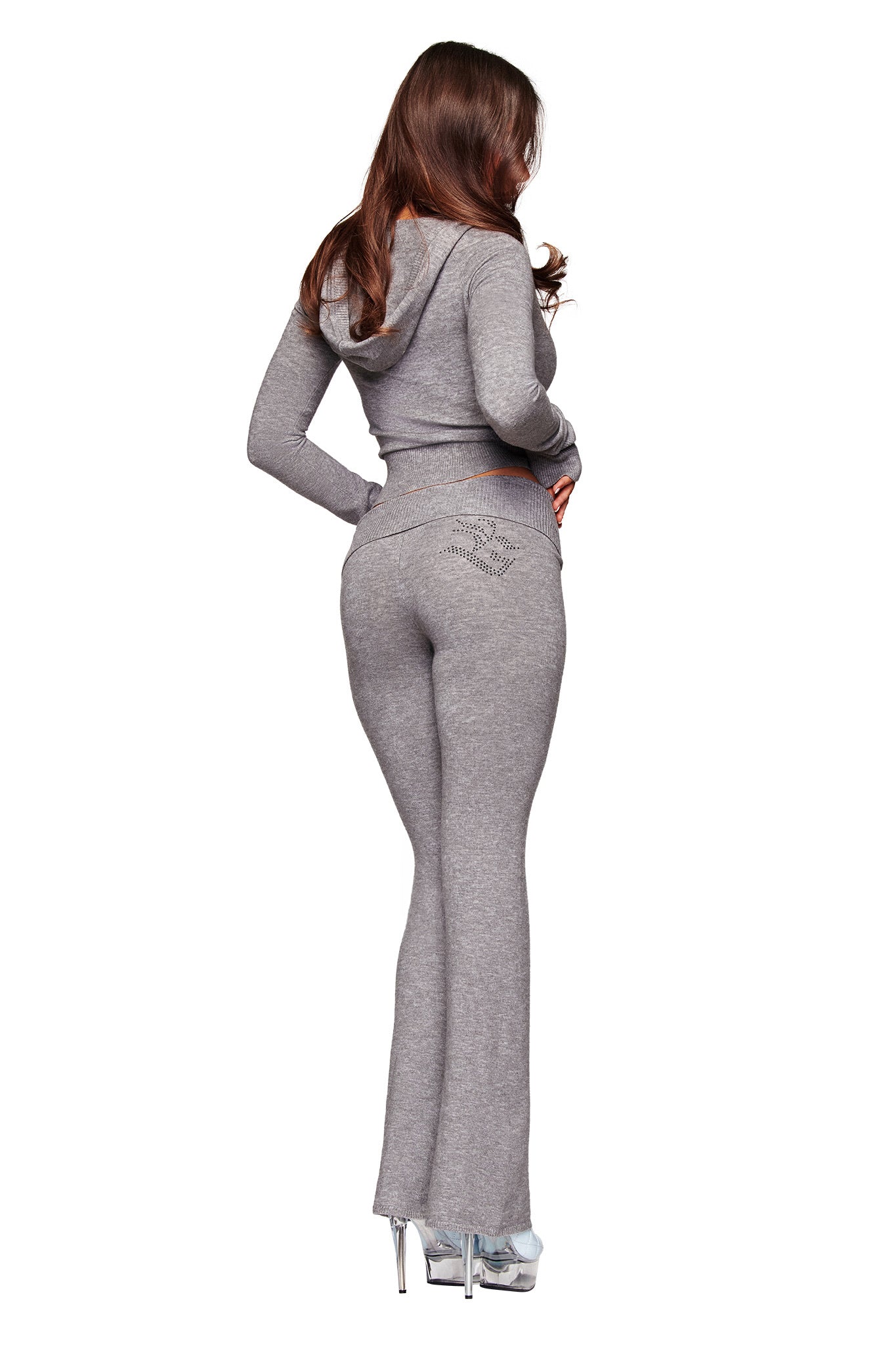 A woman with long brown hair, facing away, wears the I.AM.GIA JESSA PANT - GREY lounge tracksuit with a mid rise waist and rhinestone-detailed back pocket, paired with silver high heels. The background is white.