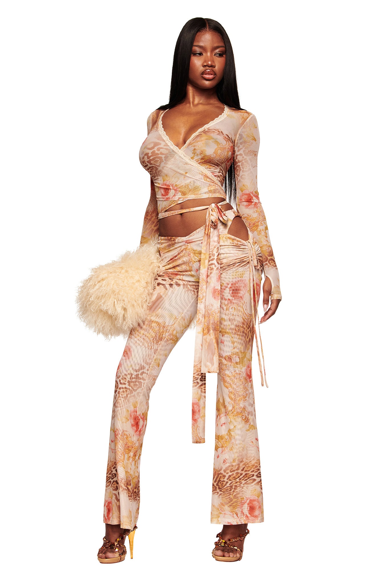 A woman wears the I.AM.GIA ERISKA PANT in Desert Rose, a pastel floral animal print with flared legs, styled with a fitted long-sleeve wrap top, high-heeled sandals, and a fluffy beige handbag.