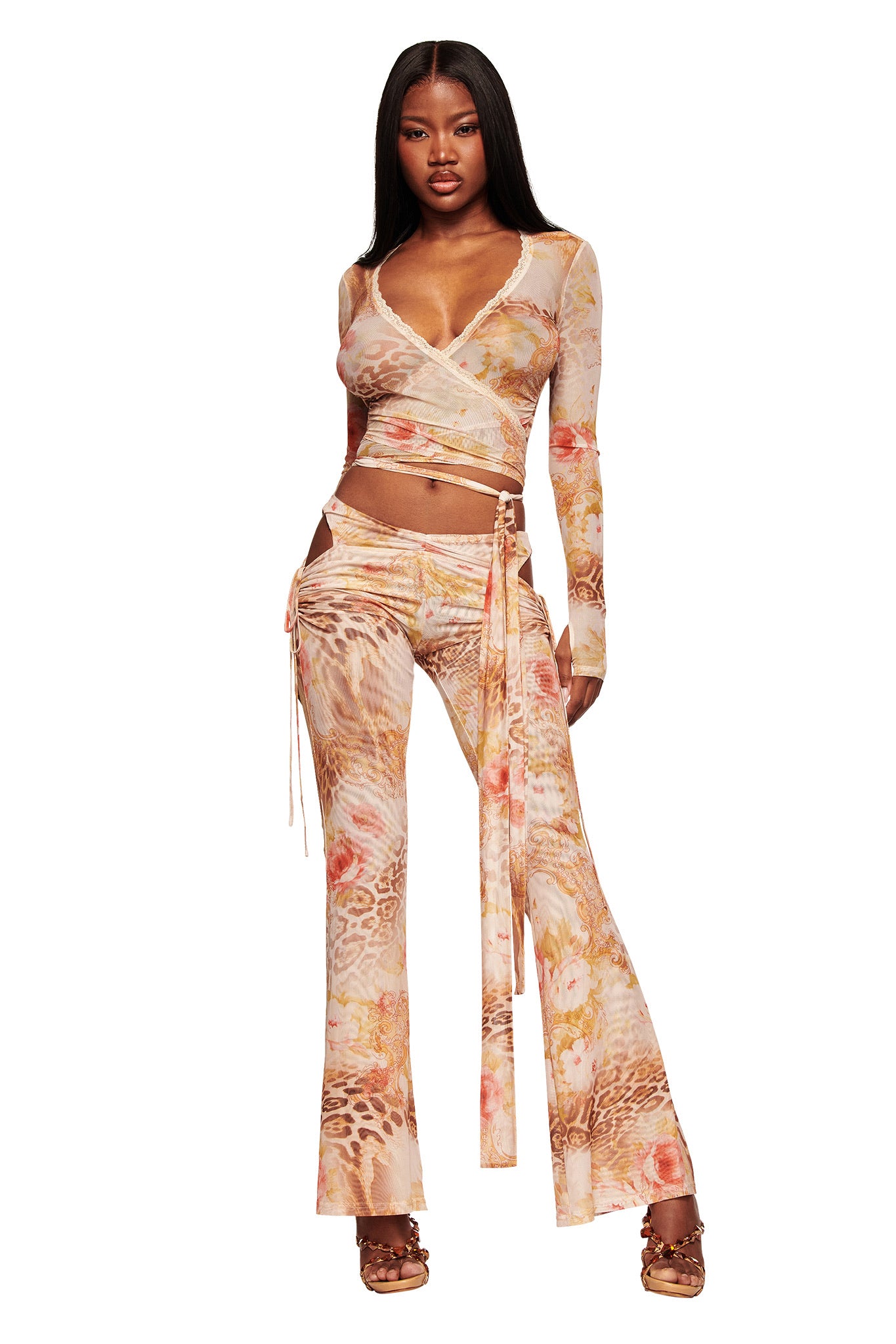 A woman models the I.AM.GIA ERISKA TOP - DESERT ROSE, a sheer mesh wrap top with an abstract animal print in pink, beige, and orange. She pairs it with matching high-waisted flared pants, brown heeled sandals, and a waist tie.