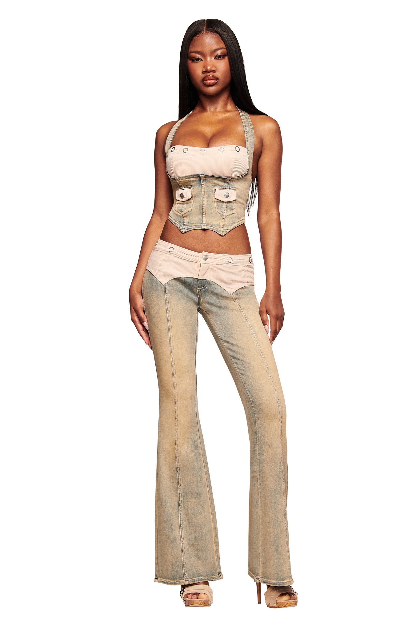 A woman faces forward in I.AM.GIA’s TOMMIE JEAN - ANTIQUE DENIM flared jeans with a mid-rise waist, paired with a light blue and beige halter top featuring buttons. She wears open-toe heels, her long straight hair completing the chic look.