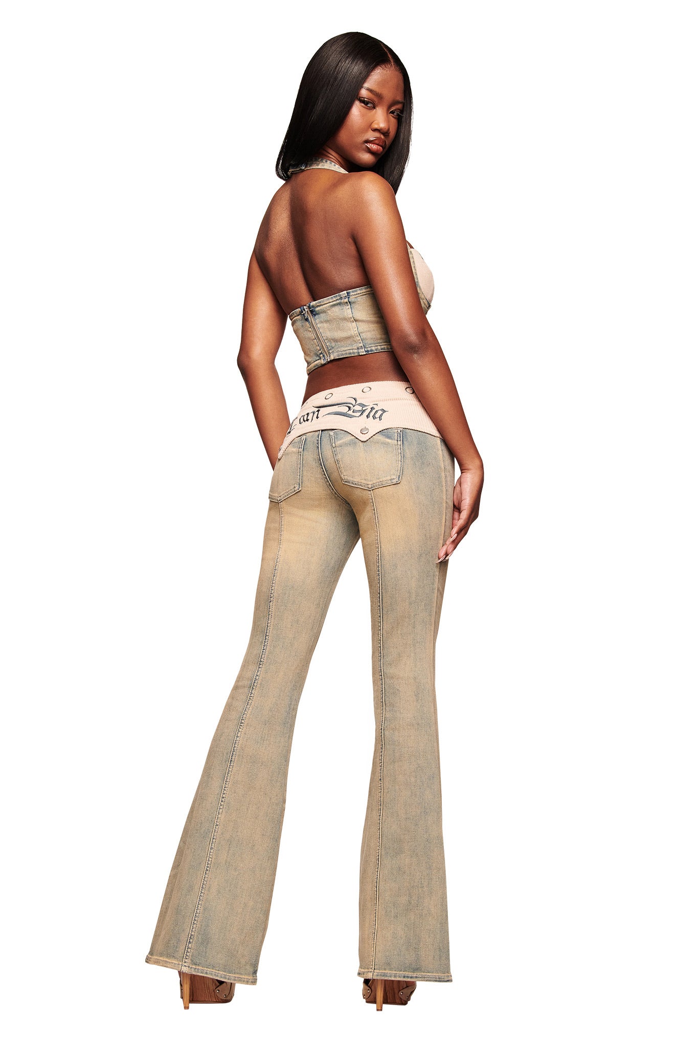 A woman with long straight hair models the I.AM.GIA TOMMIE JEAN - ANTIQUE DENIM, featuring beige flared pants with visible seams, mid rise waist, and a pink waistband. She faces away, glancing over her shoulder against a white background.