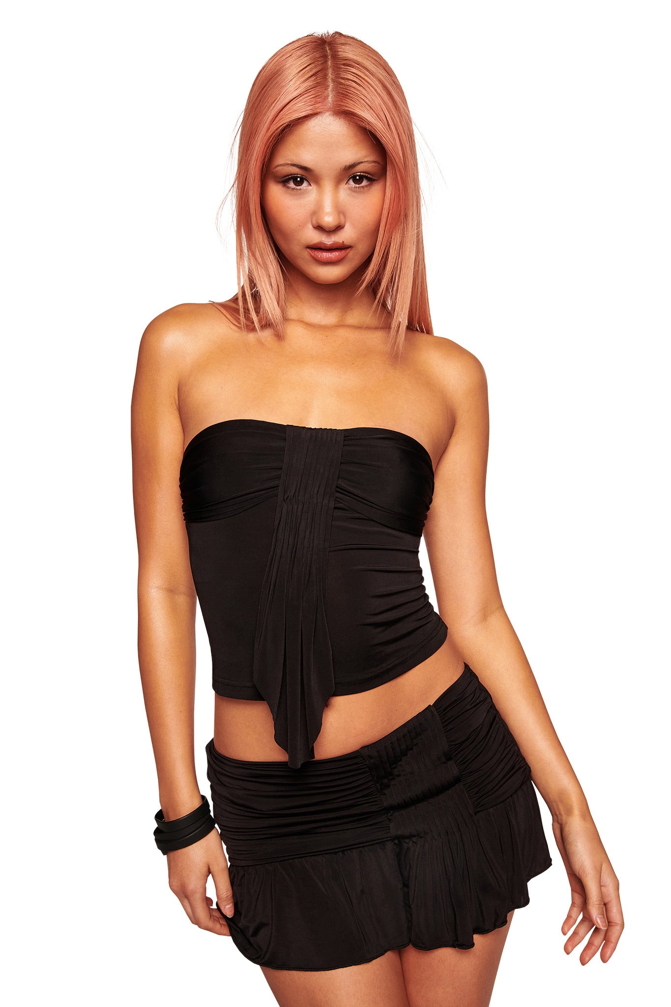 A woman with straight, shoulder-length pink hair wears the I.AM.GIA ELYSIA BOOB TUBE in black, paired with a matching short slinky knit skirt. She stands against a white background, looking confidently at the camera.