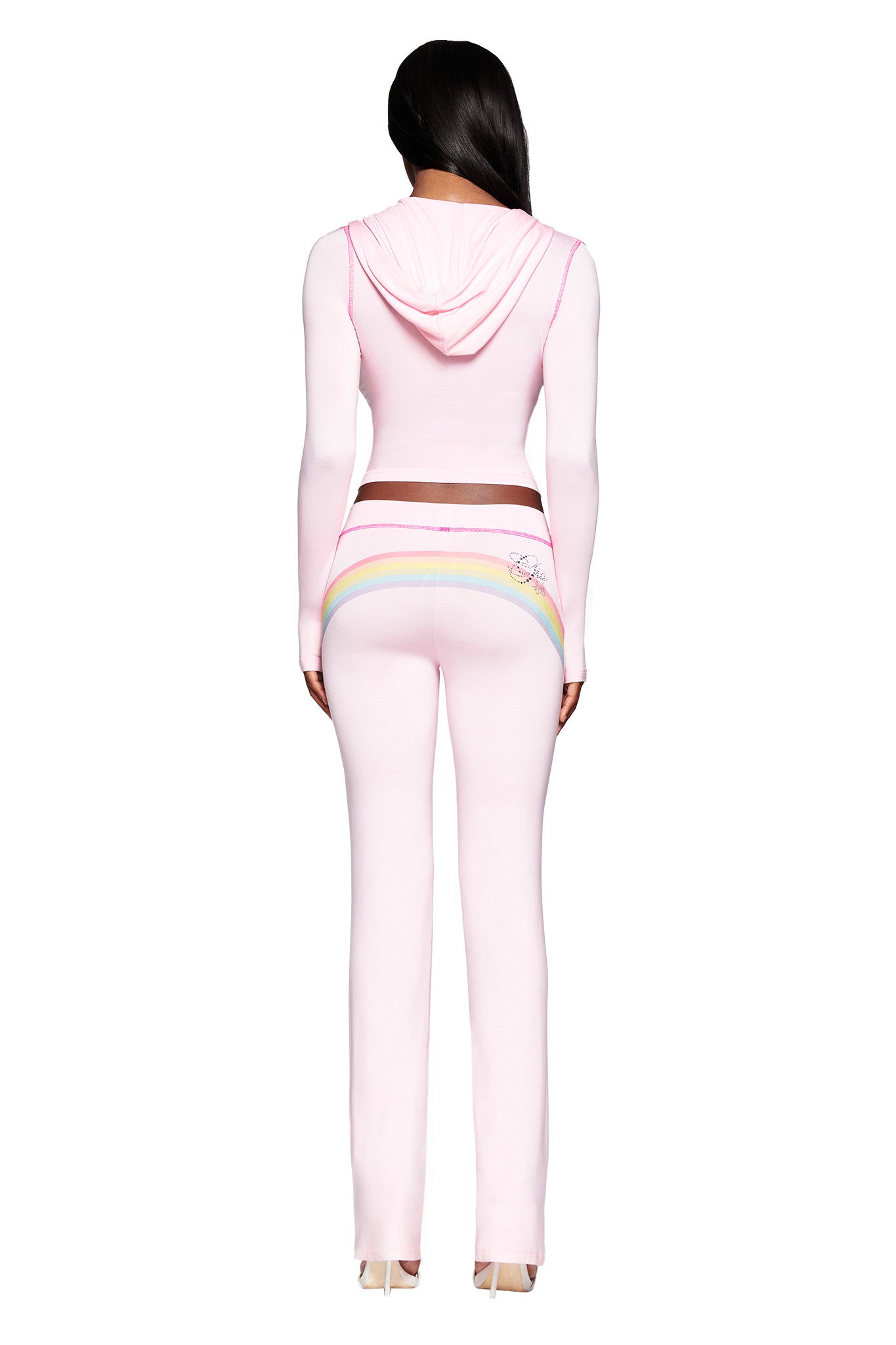 A woman faces away in the I.AM.GIA FAFI BLARE HOODIE - BABY PINK and matching fitted pants, featuring pastel rainbow script details. The light pink, body-con set stands out against a plain white background and is styled with high heels.