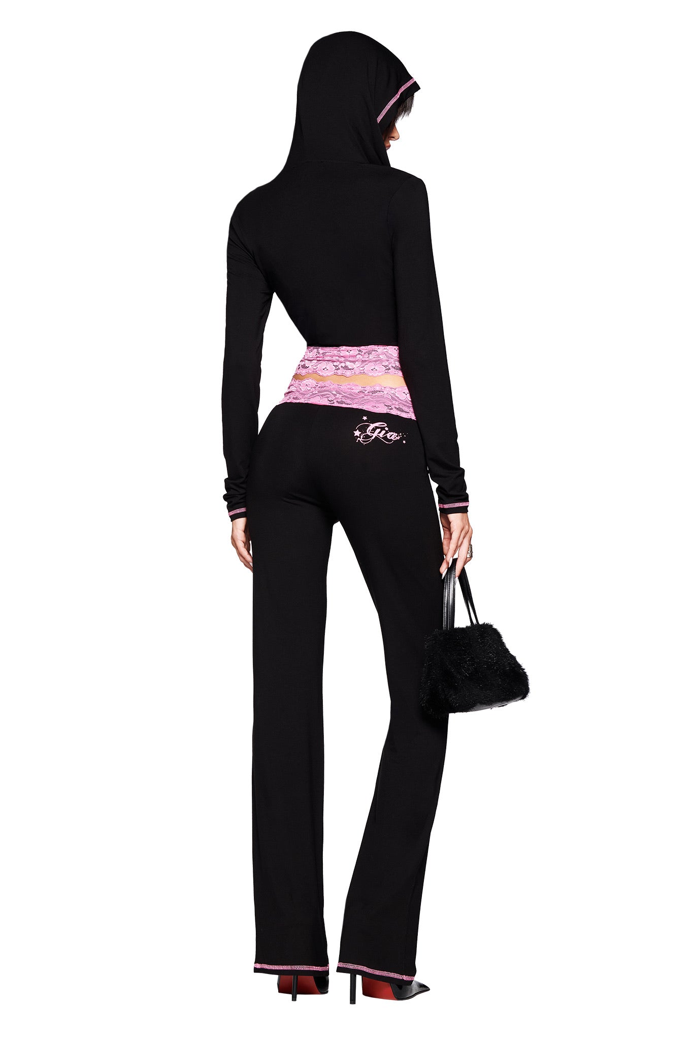 A woman stands with her back to the camera wearing I.AM.GIA's TARSHA PANT in black/pink, styled with a black hoodie, pink waistband, black fuzzy handbag, and high heels.