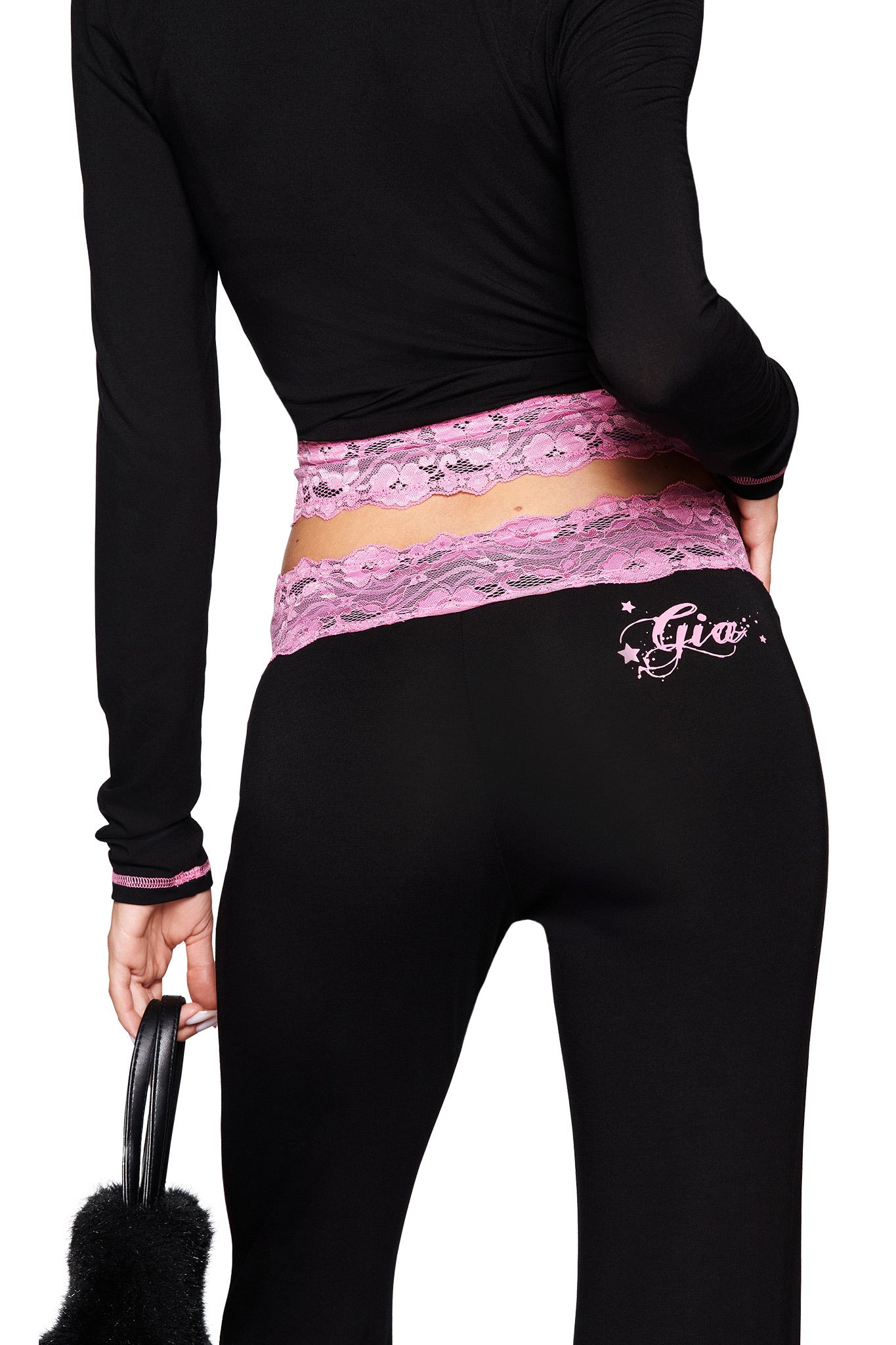 Wearing a black long-sleeve top and I.AM.GIA TARSHA PANT in black/pink with a pink lace mid-rise waist, "Gia" with stars on the back, and holding a black furry accessory.