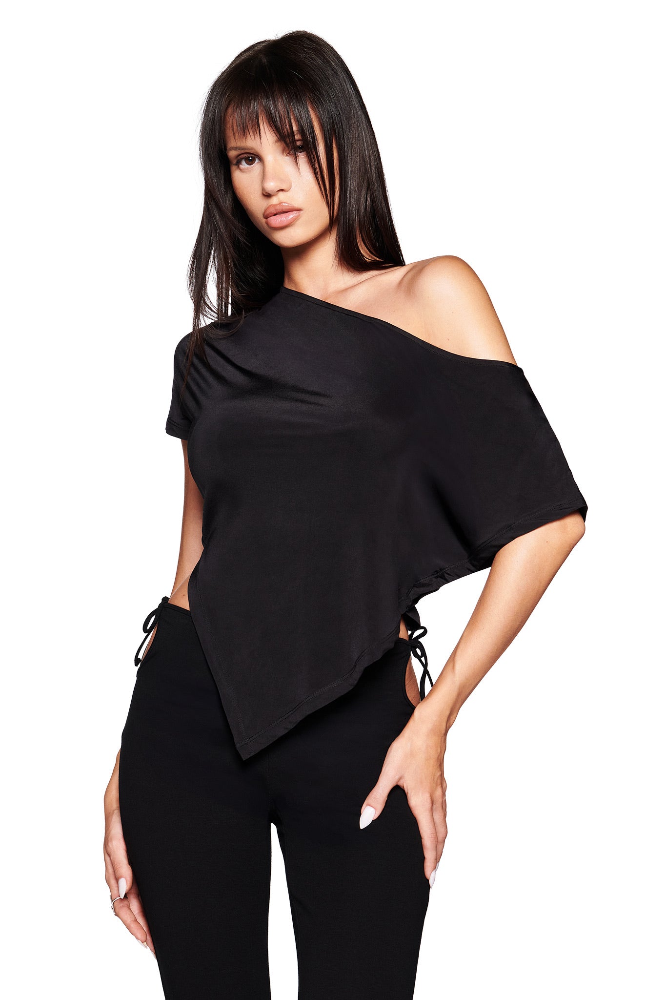A woman in the I.AM.GIA JOVIE TOP - BLACK, featuring an asymmetrical one-shoulder design with side ties and hem, is paired with black pants and stands against a white background.
