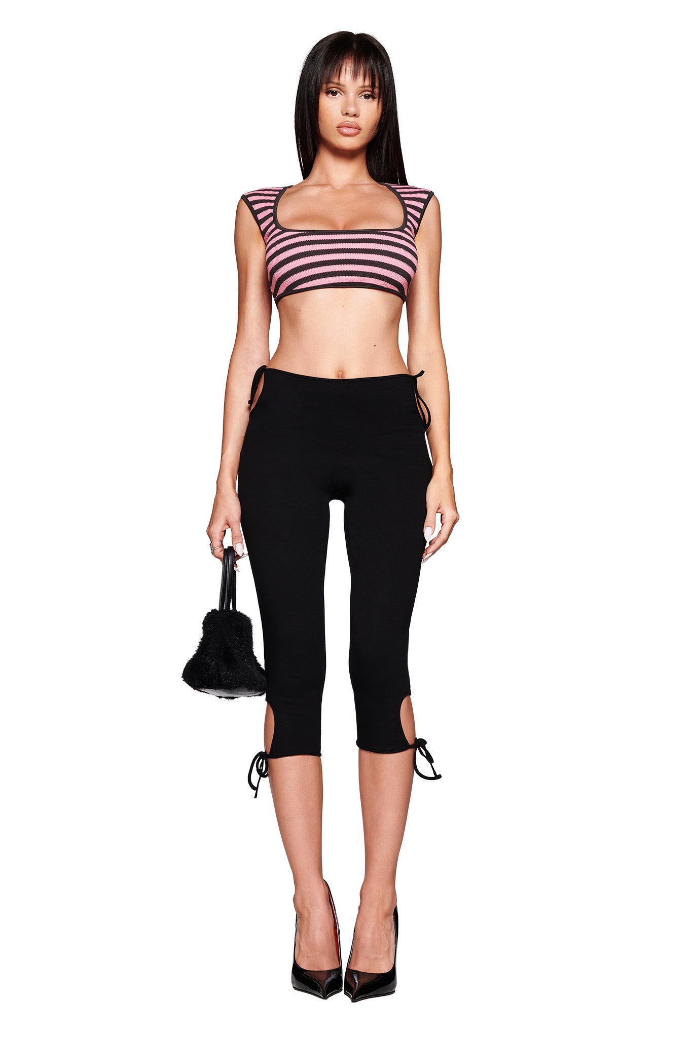 A woman faces forward, wearing I.AM.GIA's YUNA CAPRI - BLACK lace-up capri pants, a pink and black striped body-con crop top, black high heels, holding a small black fuzzy handbag. She has straight long hair with bangs.