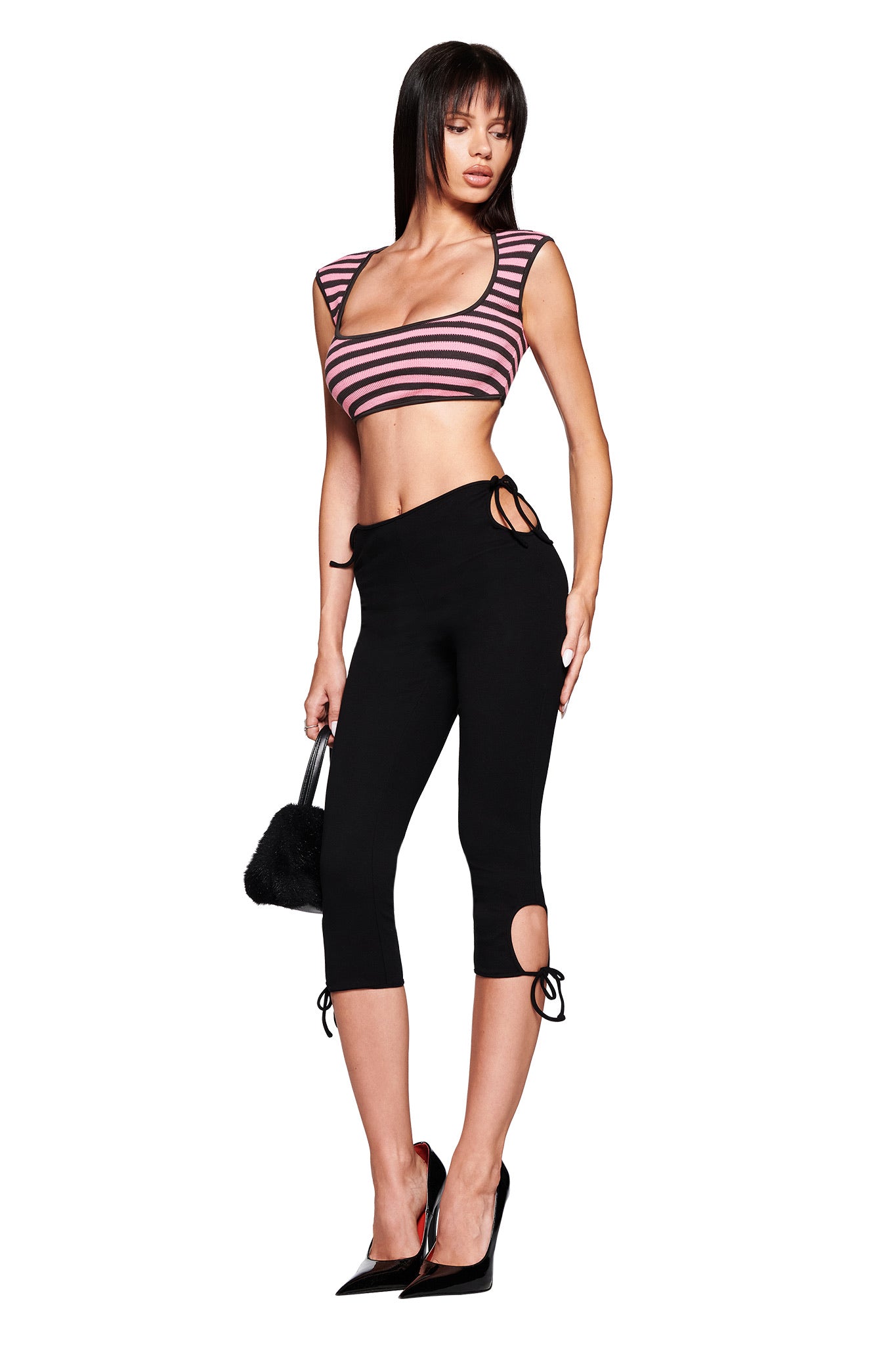 A woman poses against a white background wearing I.AM.GIA's YUNA CAPRI - BLACK in a body-con fit with side ties, paired with a pink and black striped crop top, black high heels, and holding a small black handbag.