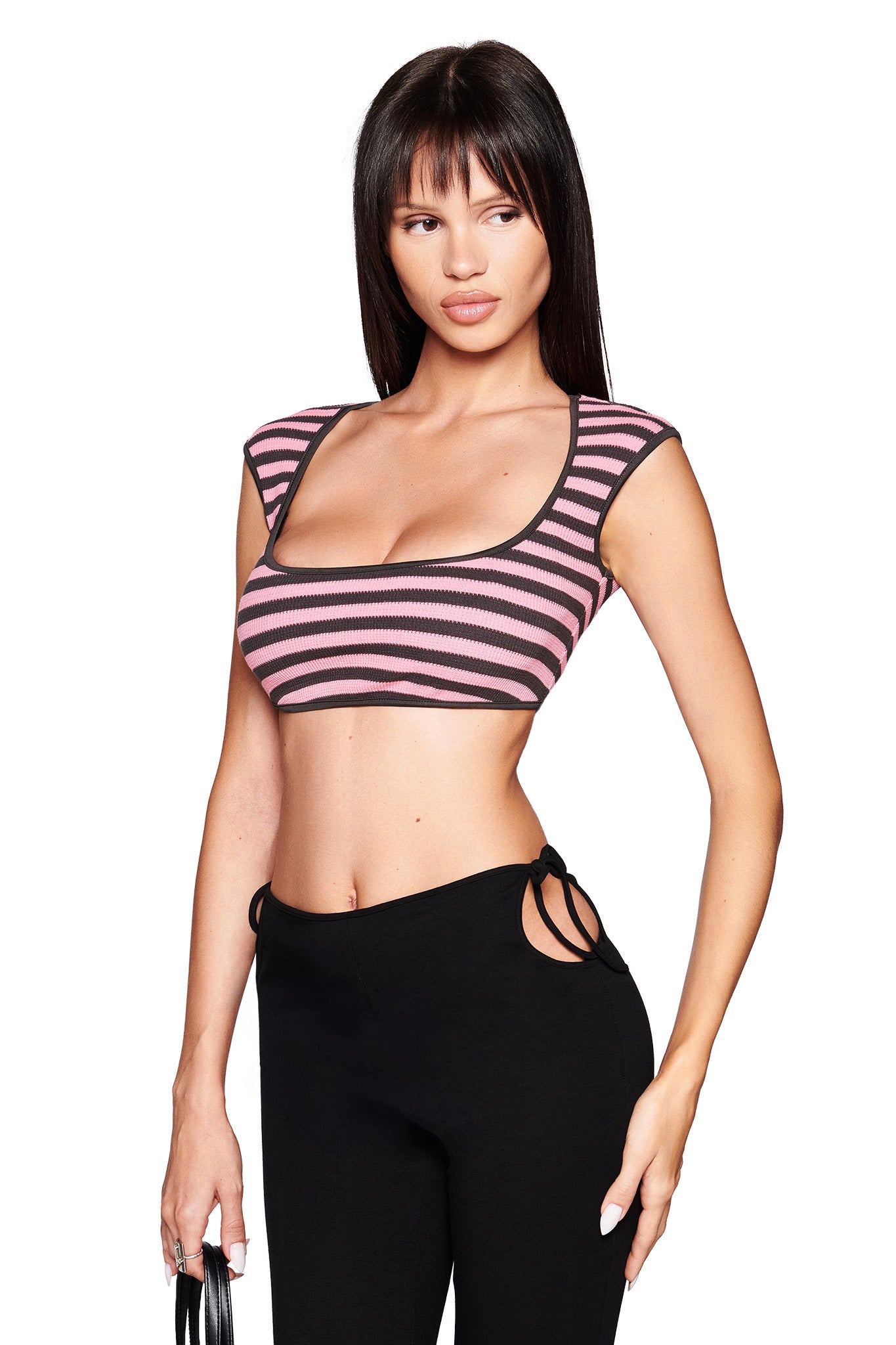 A woman with straight dark hair wears the I.AM.GIA RICA TOP in pink/grey and black high-waisted pants with side cutouts, standing against a white background and holding a black strap.