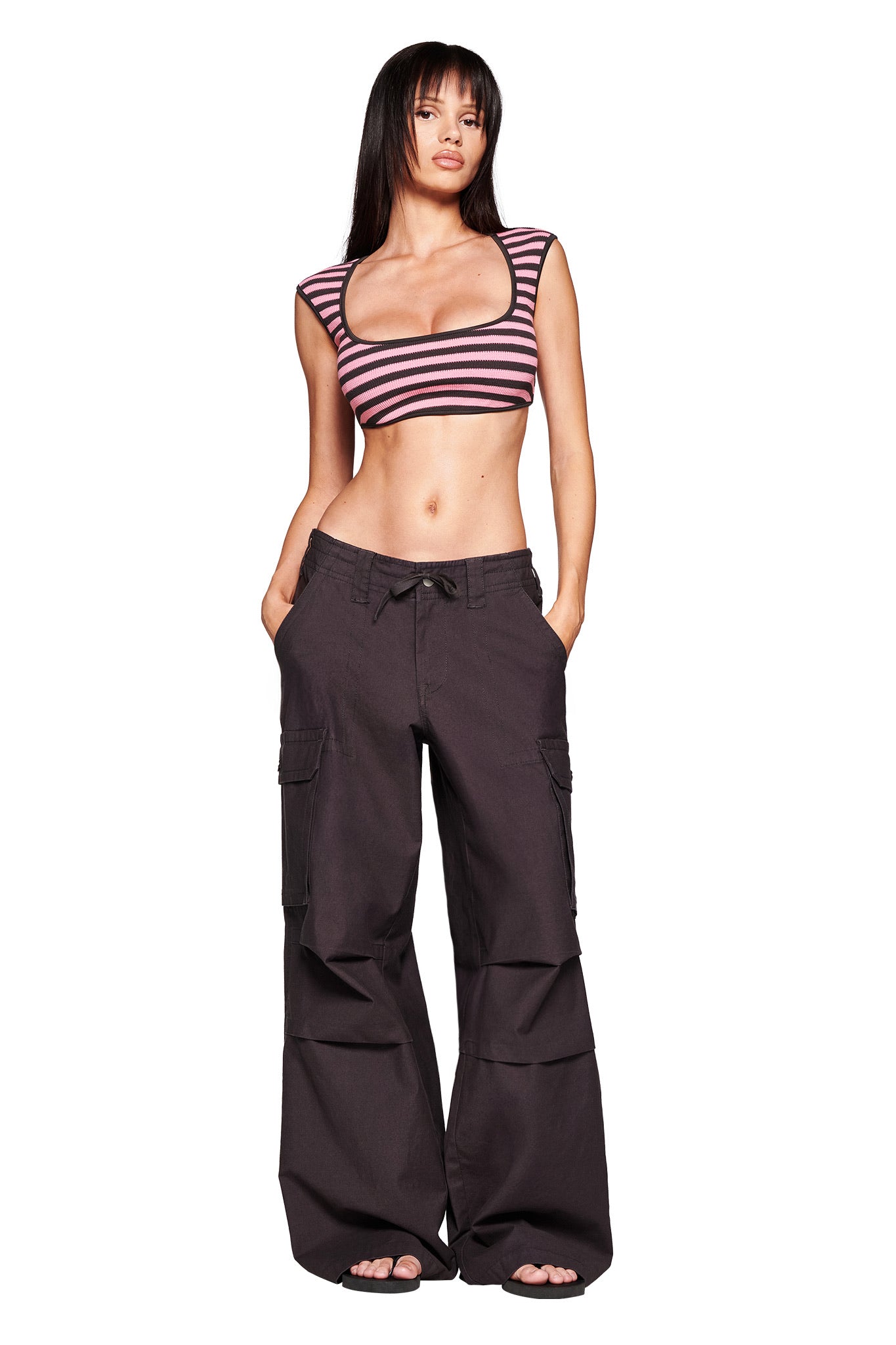A woman with long straight hair and bangs stands barefoot against a white background, wearing a pink and black striped crop top and I.AM.GIA JAXXON PANT - GREY cargo pants with an oversized fit, hands in her pockets.