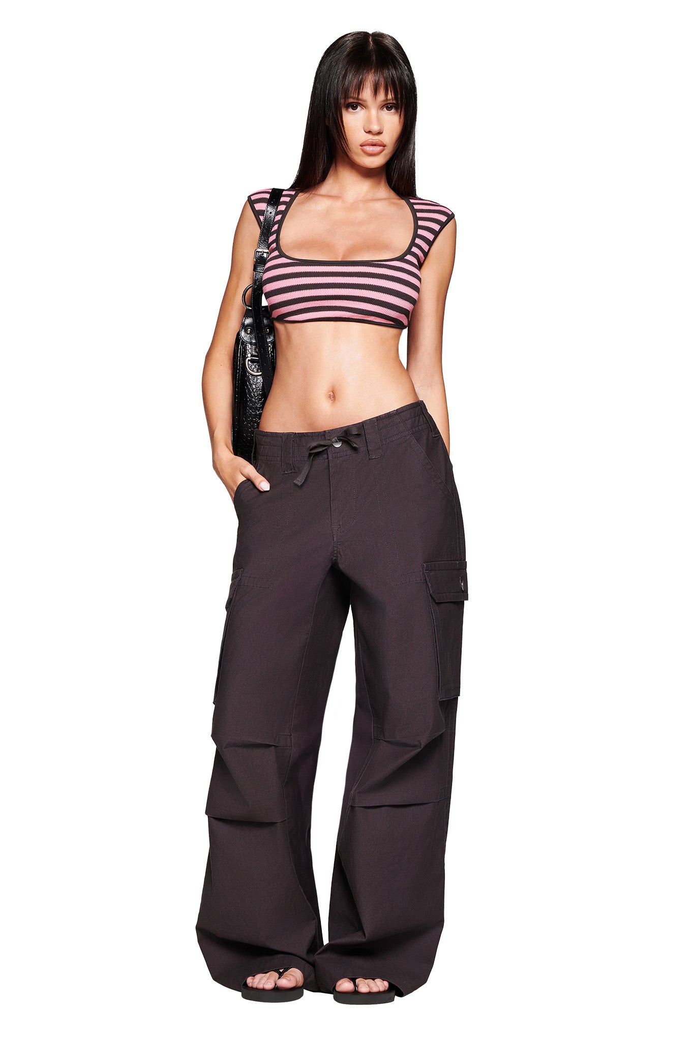 A woman stands confidently in the I.AM.GIA RICA TOP in pink and grey, paired with loose black cargo pants, sandals, a black shoulder bag, and her long straight dark hair styled with bangs.