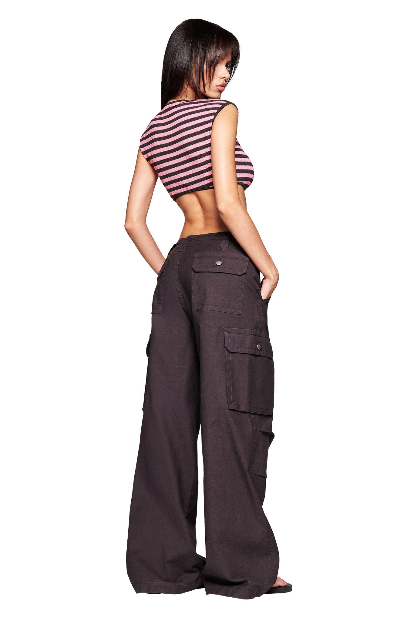 A woman with straight dark hair stands facing away, wearing the I.AM.GIA RICA TOP in pink and grey—a striped waffle knit sleeveless crop tank—paired with loose high-waisted black cargo pants with pockets, hands in her pockets, against a white background.
